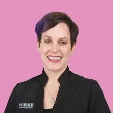 A woman with short dark hair smiling, wearing a black jacket against a pink background.