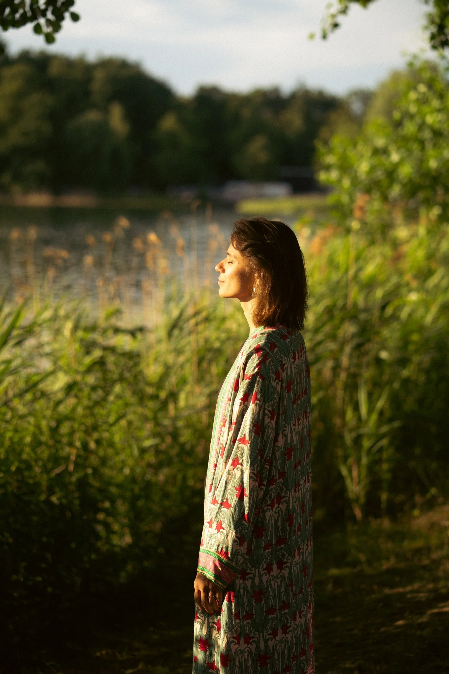Woman standing in profile with eyes closed, illuminated by sunlight, near a lake and greenery in the background.