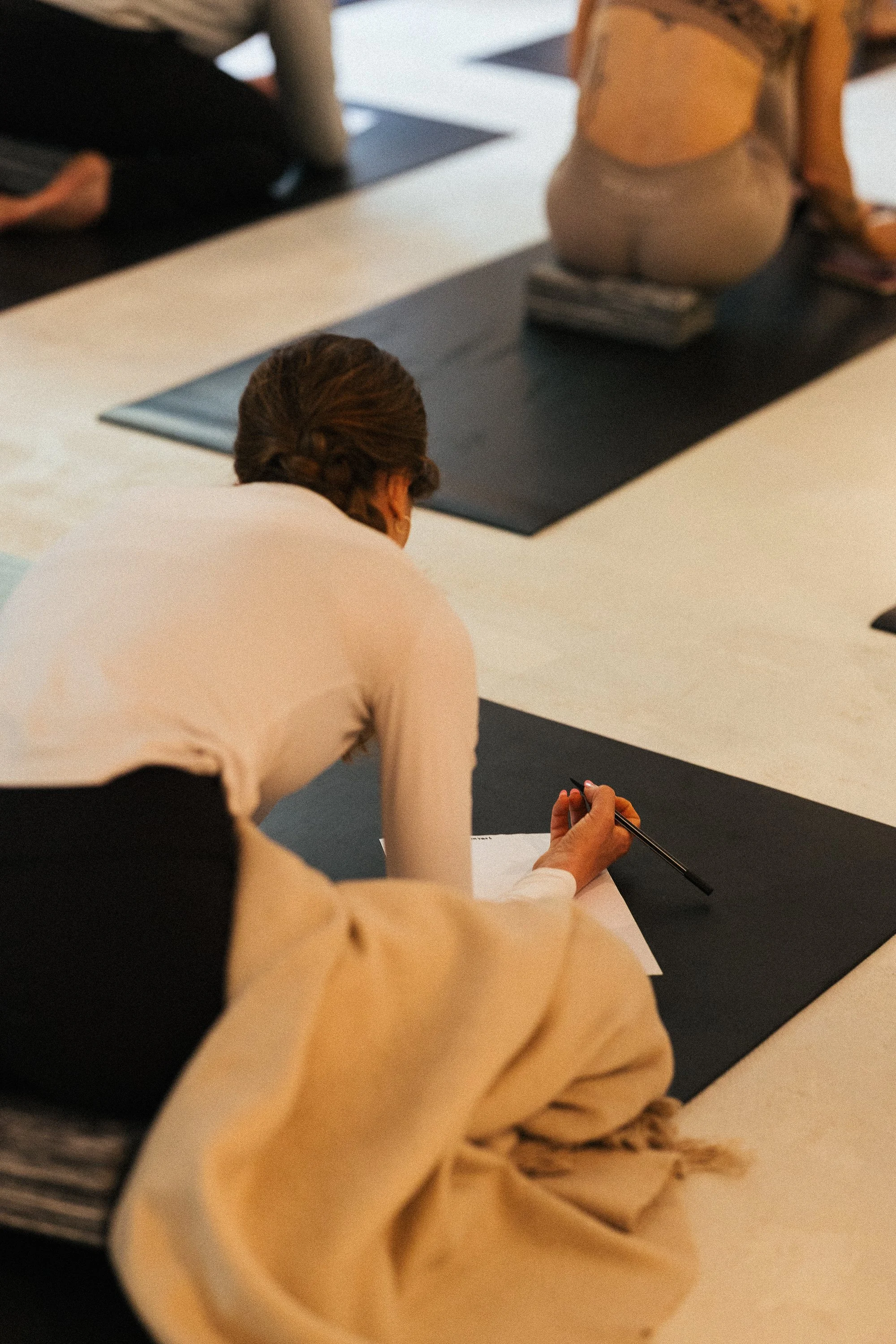 A woman with brown hair sitting on a black yoga mat on a large white mat in a yoga class, holding a pen and writing on a piece of paper. Other people are seated on mats in the background.