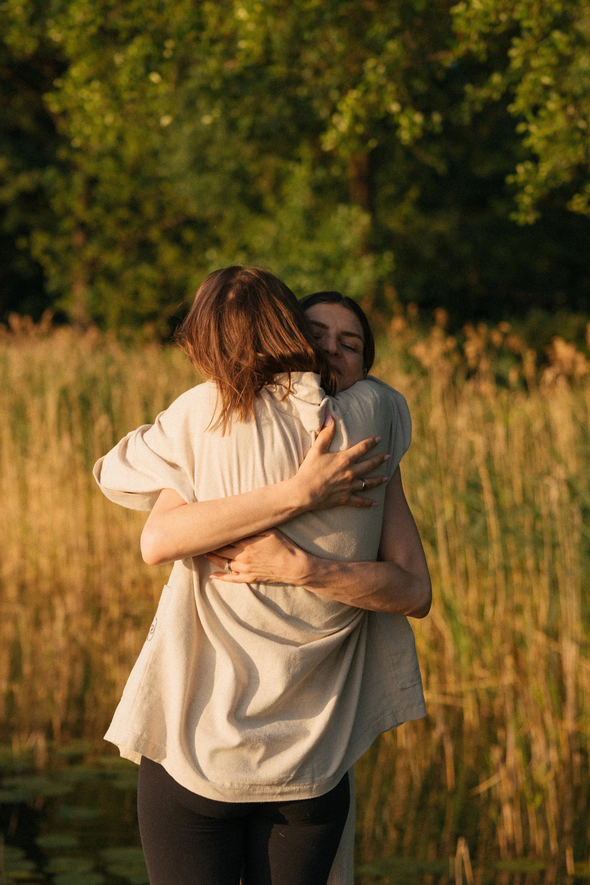 Two women hugging outdoors in a natural setting with trees and tall grass, bathed in warm sunlight.