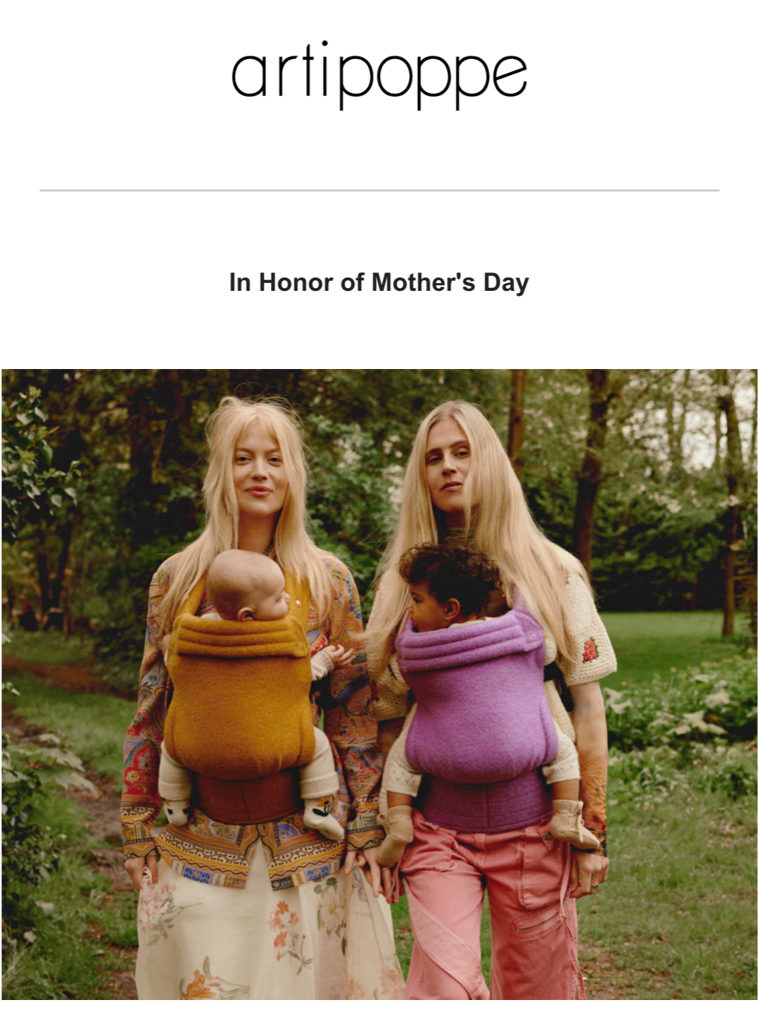 Two women walking outdoors in a park, each carrying a baby in a colorful wrap carrier, surrounded by greenery.