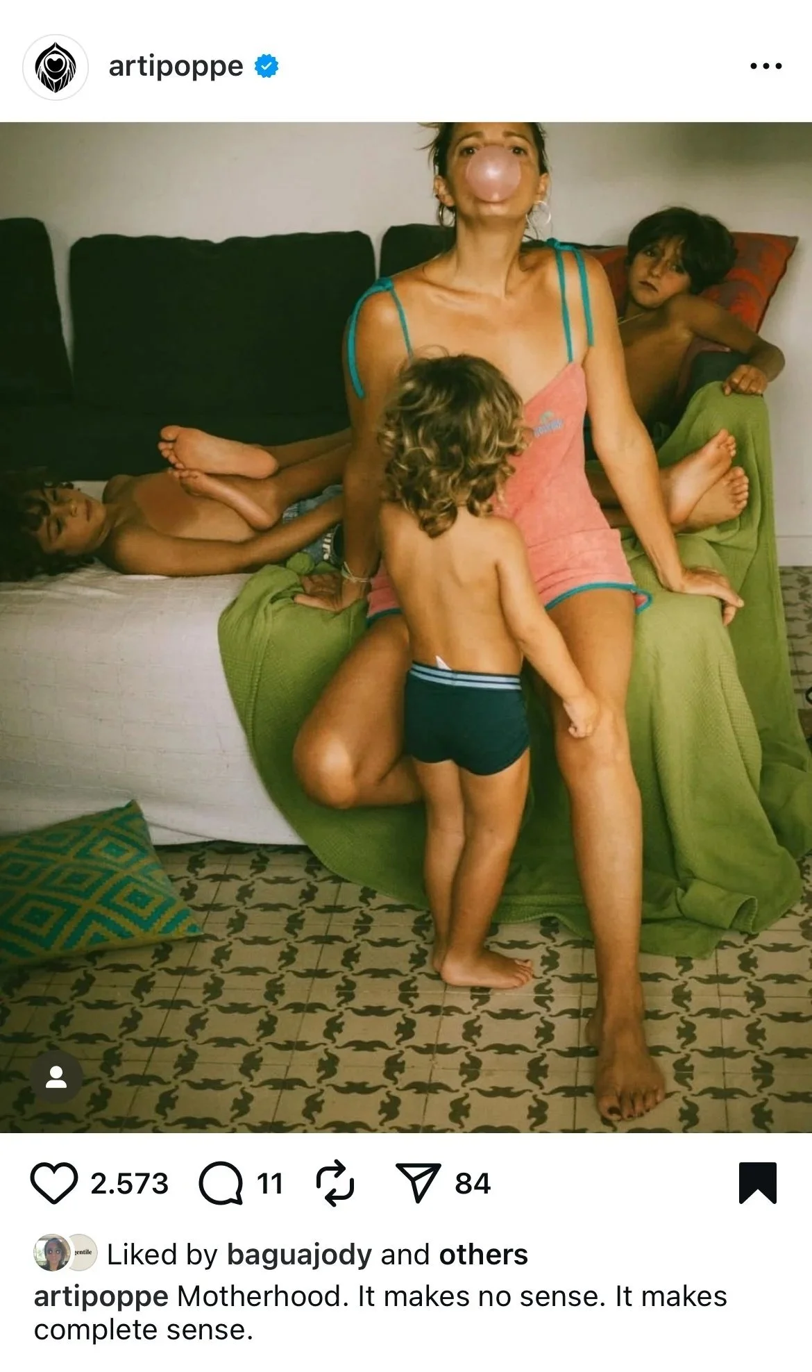 A woman with a large nose filter sitting on a sofa with three children around her, all in underwear. One child is lying on the sofa, another is standing next to her, and the third is also lying on the sofa. The room has a patterned floor, a green bla