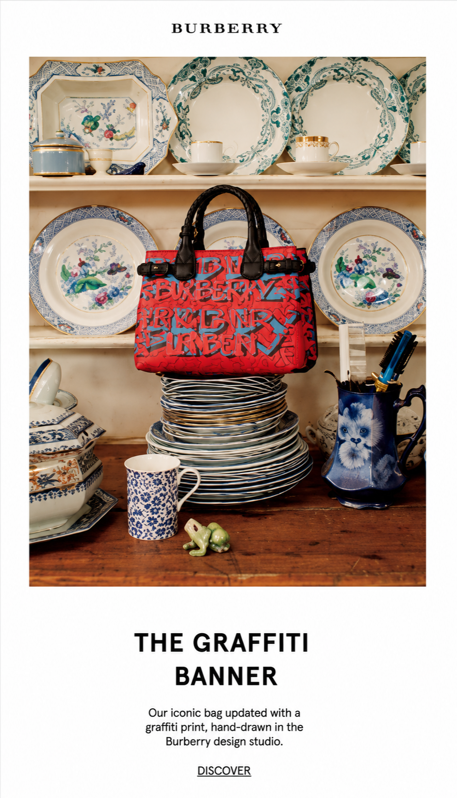 A display of blue and white patterned china plates and bowls on a wooden surface, with a red Burberry bag with graffiti print hanging among the plates and bowls, a blue and white mug, a small green frog figurine, and a blue pitcher with a lion image 