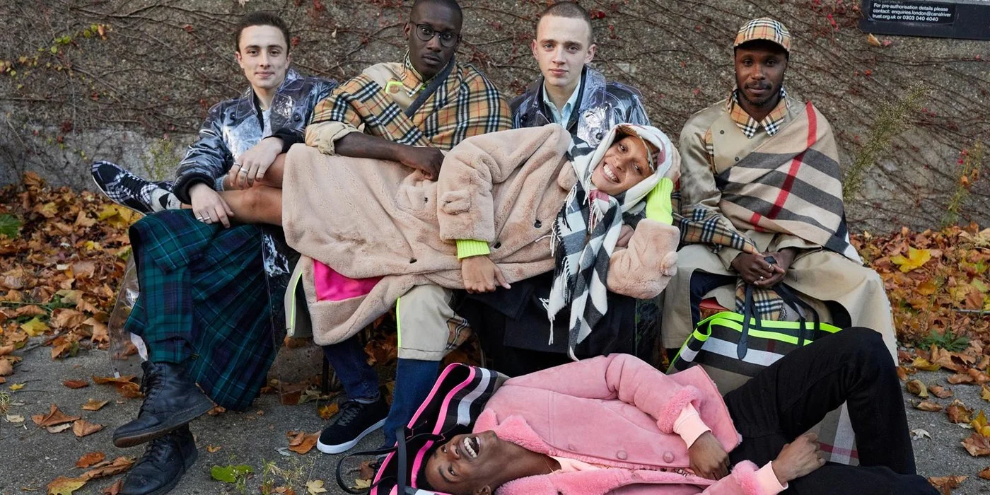 Six young models in Burberry trench coats sitting on and lying on the ground with fallen autumn leaves, some wearing plaid scarves and jackets, in front of a brick wall. The Giant Burberry runway bag is in the shot