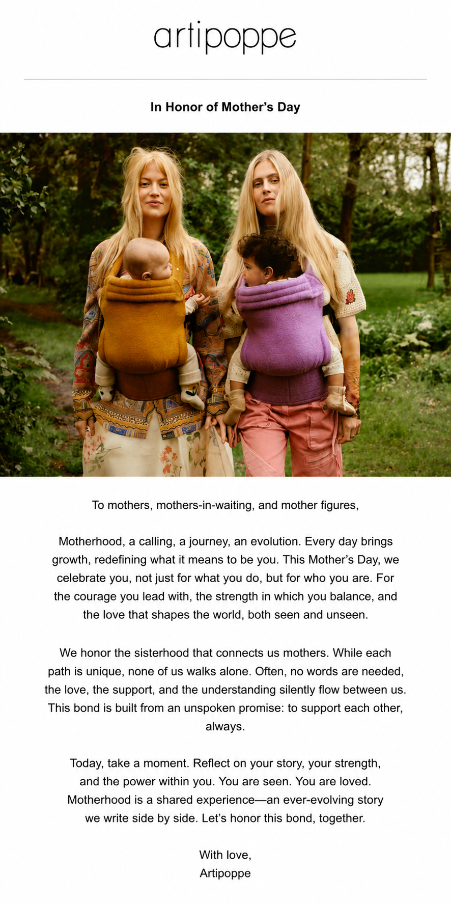 Two stylish blonde women walking outdoors in a park, each carrying a baby in a colorful Artipoppe carrier, surrounded by greenery.