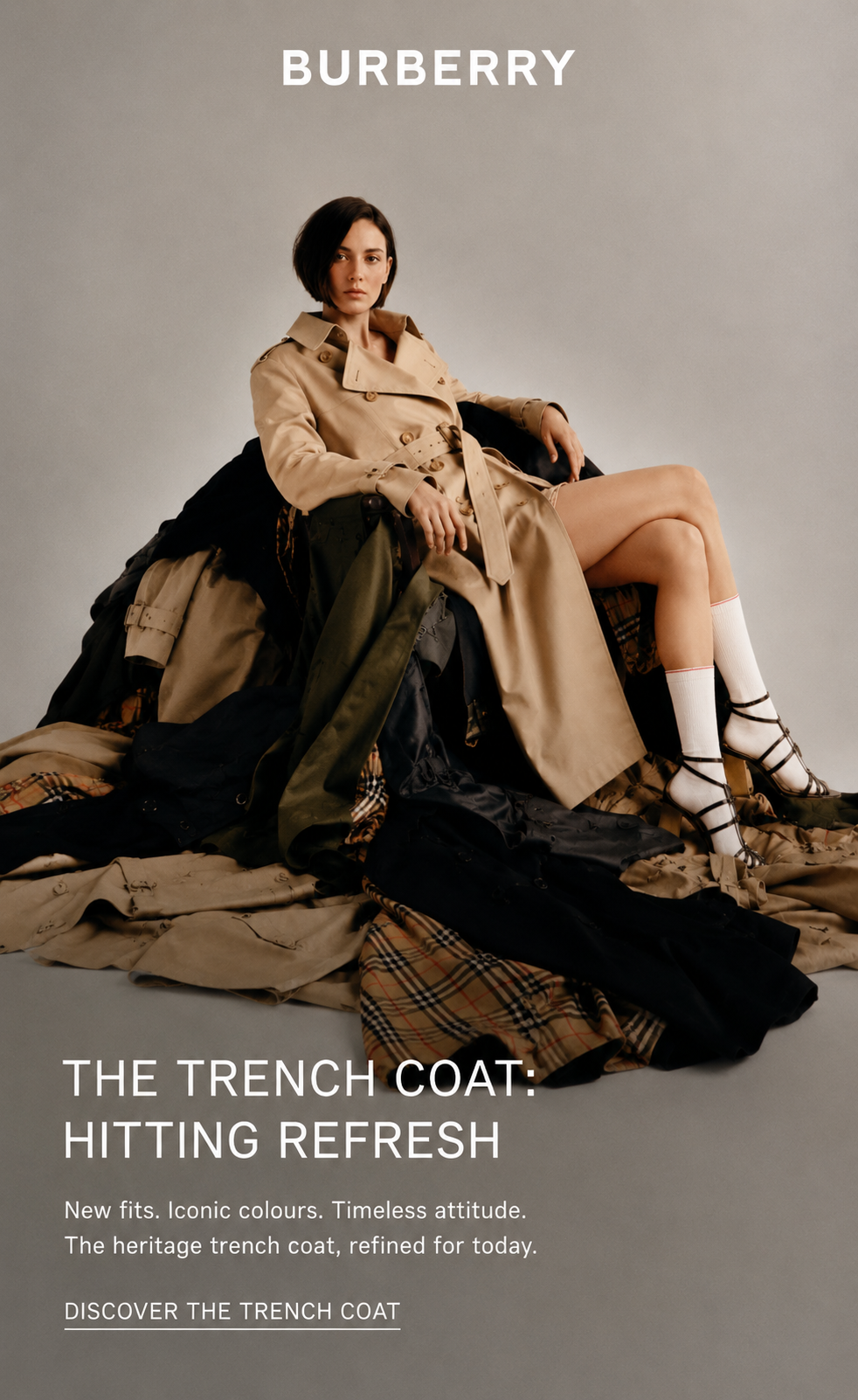 A woman seated on a pile of trench coats, wearing a beige Burberry trench coat and white socks with black strappy sandals. The background is neutral gray, with white text promoting Burberry's trench coat refresh collection.