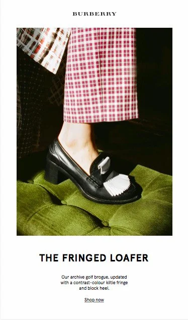 Close-up of a model wearing a pair of black Burberry fringe loafers with white fringe details, standing on a green velvet surface, with pink and checkered patterned pants.