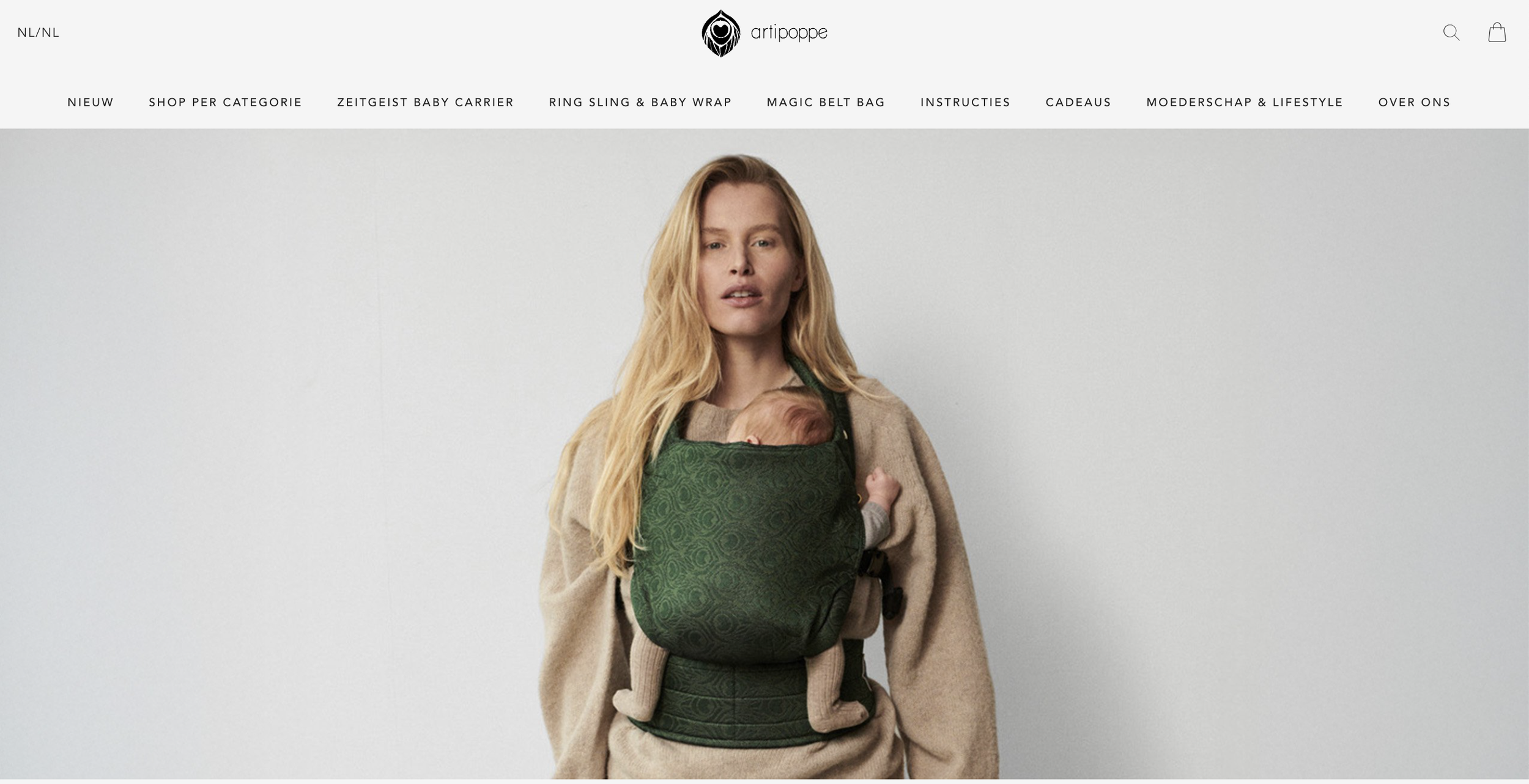 A woman with long blonde hair wears a beige coat and carries a baby in a green baby carrier on her chest.