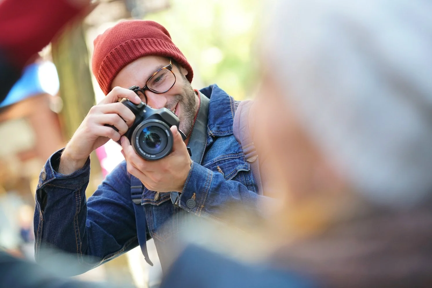 Man in a red knit hat, holding a camera up in front of his smiling face as he takes a photo of someone