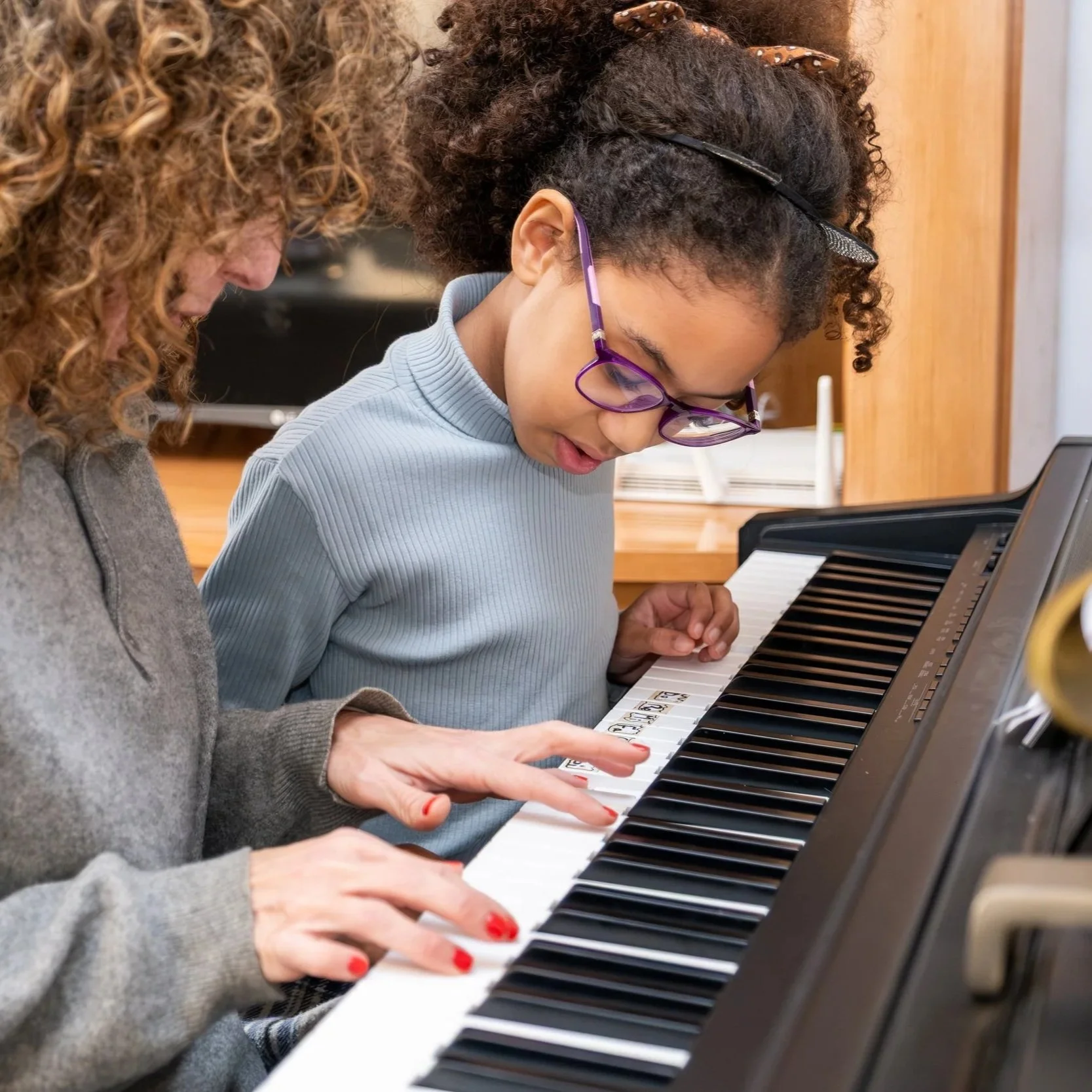 A young child is sitting in front of a piano learning how to play. Their piano teacher is sitting beside them with their hands on the piano keys.