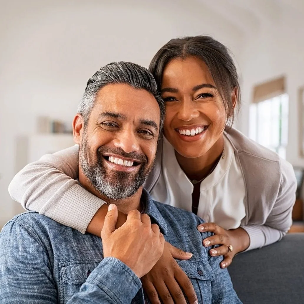 Portrait of multiethnic couple embracing and looking at camera sitting on sofa. Smiling woman hugging man sitting on couch from behind at home. Happy mature mixed race couple laughing at home.