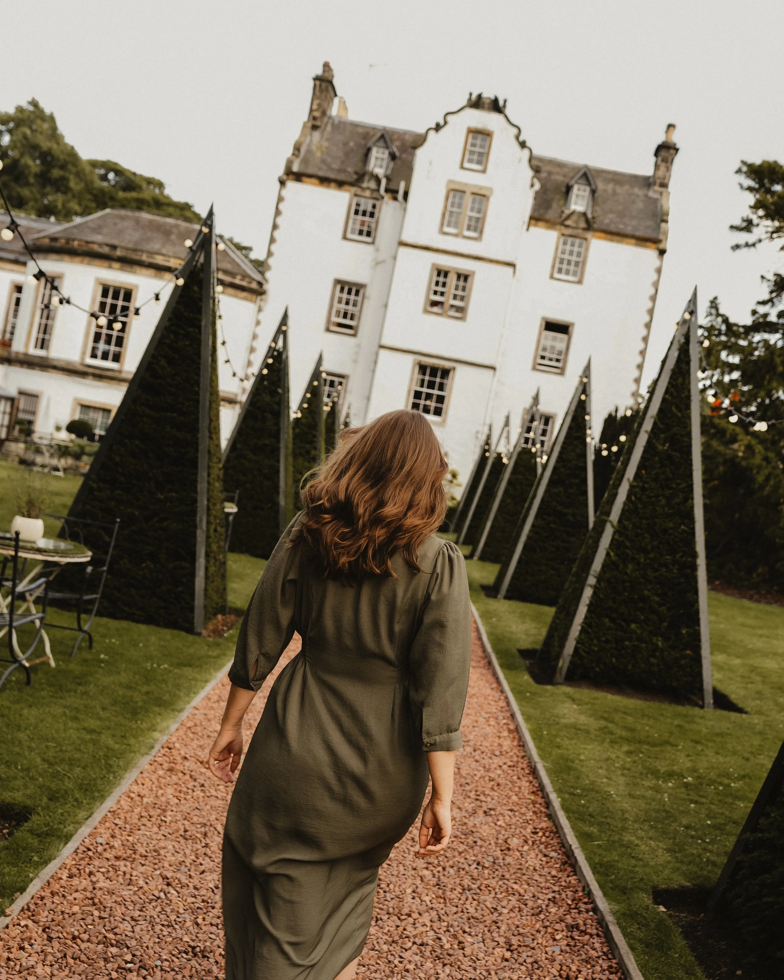 A woman walking on a gravel path lined with tall, triangular, dark green bushes leading to a historic white mansion with multiple chimneys and windows.