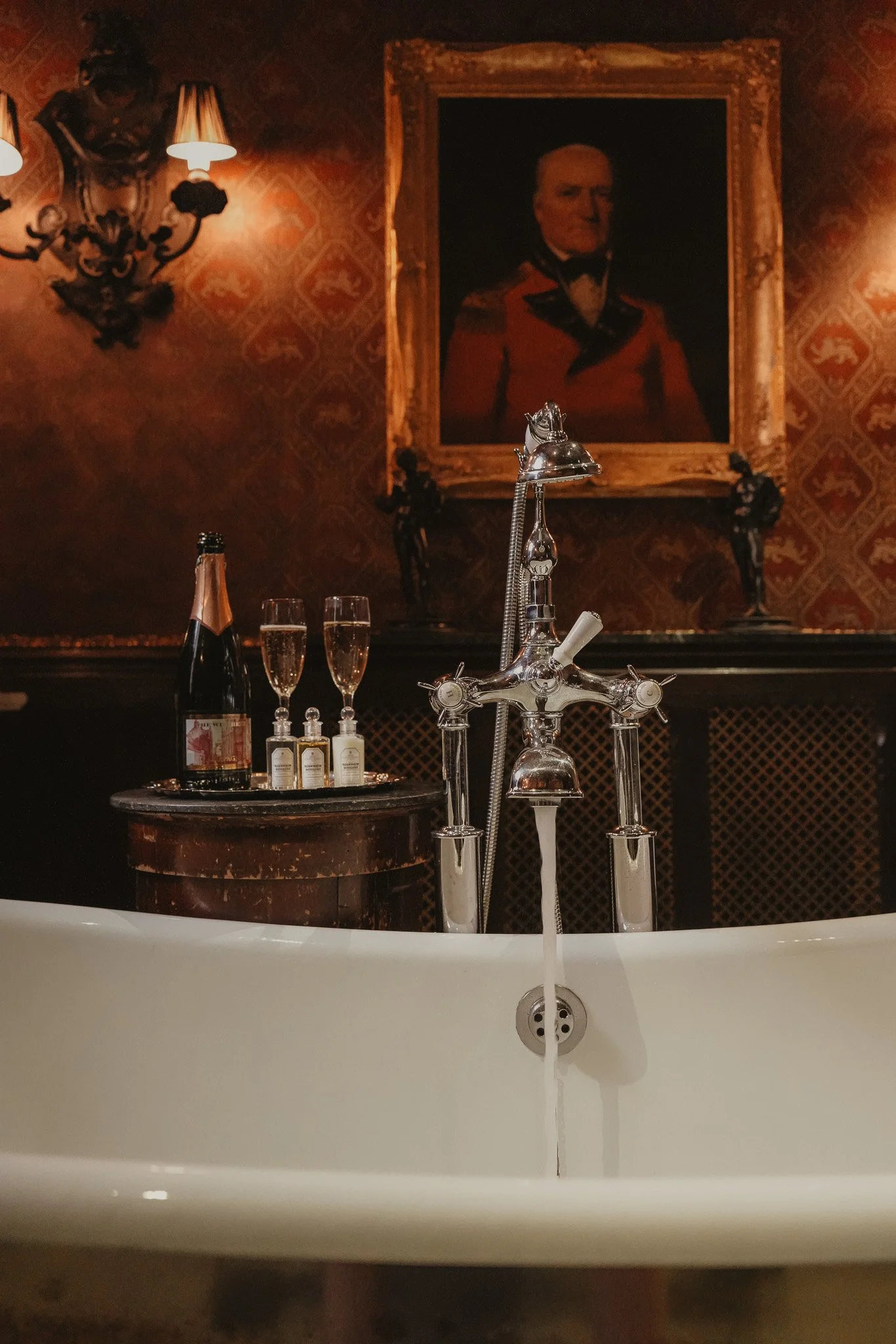A vintage-style bathtub with a shiny chrome faucet and a white interior. On a small round table behind the tub, there are two bottles of champagne and two glasses filled with champagne. The background features a wall with ornate dark wallpaper, a large framed portrait of a man in a red military uniform, and a decorative wall sconce with lit lamps.