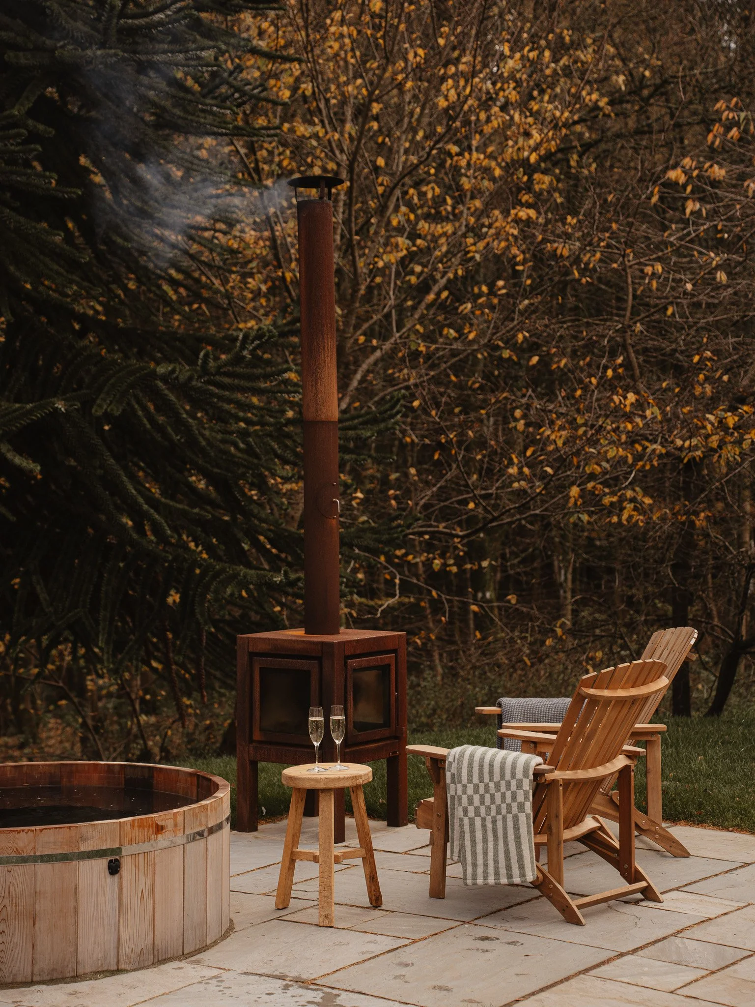 An outdoor patio scene with Adirondack chairs, a small wooden table with two glasses of champagne, a wood stove with a visible chimney emitting smoke, a hot tub, and autumn trees in the background.
