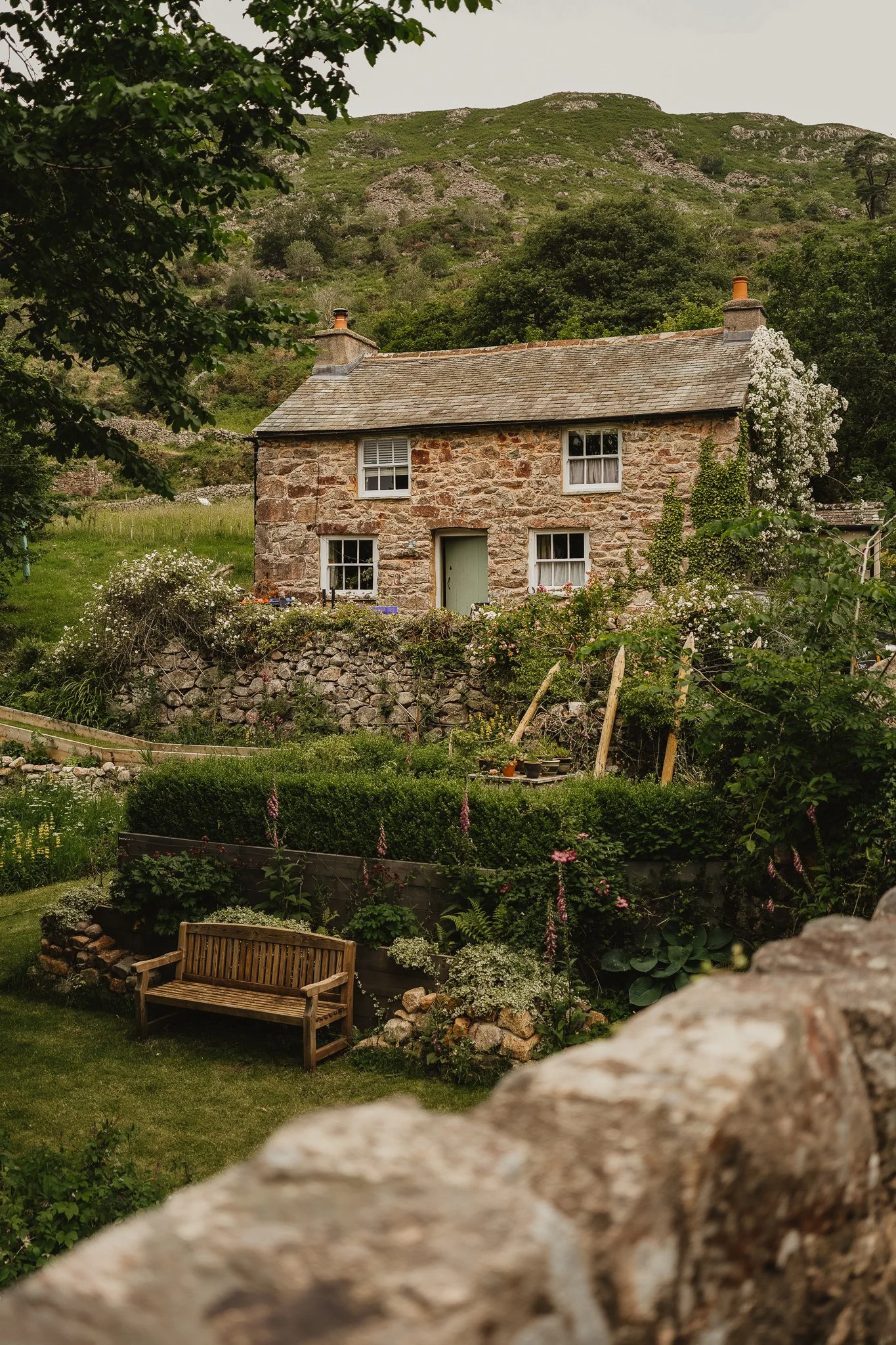 A rustic stone house with three windows and a light green door, surrounded by greenery, flowering plants, and a garden with a wooden bench in a lush countryside setting with hills in the background.