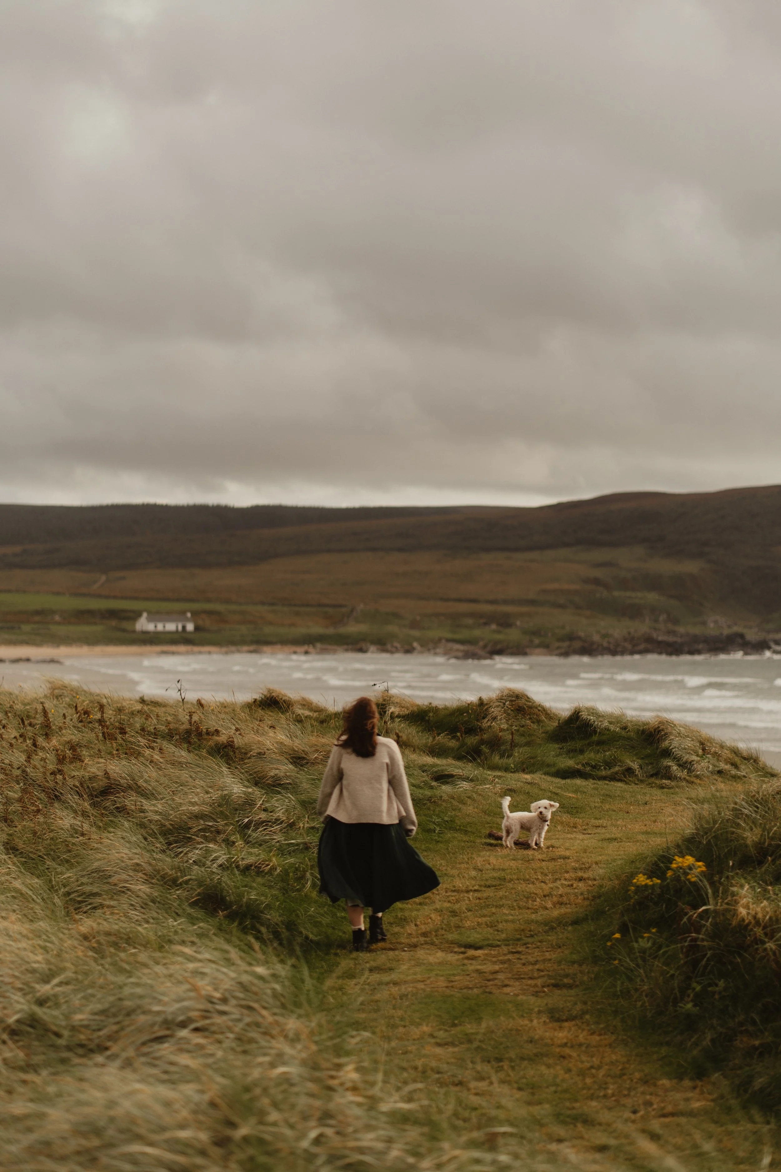A woman walking along a grassy coastal path with a dog, overlooking the ocean with rolling hills and a cloudy sky in the background.