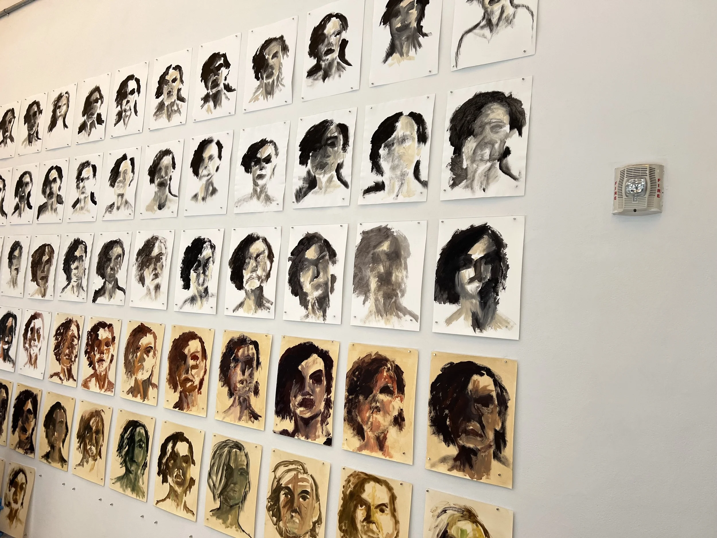 A wall display of multiple portrait paintings of a woman's face, arranged in rows, each painted with bold brushstrokes in various color palettes.