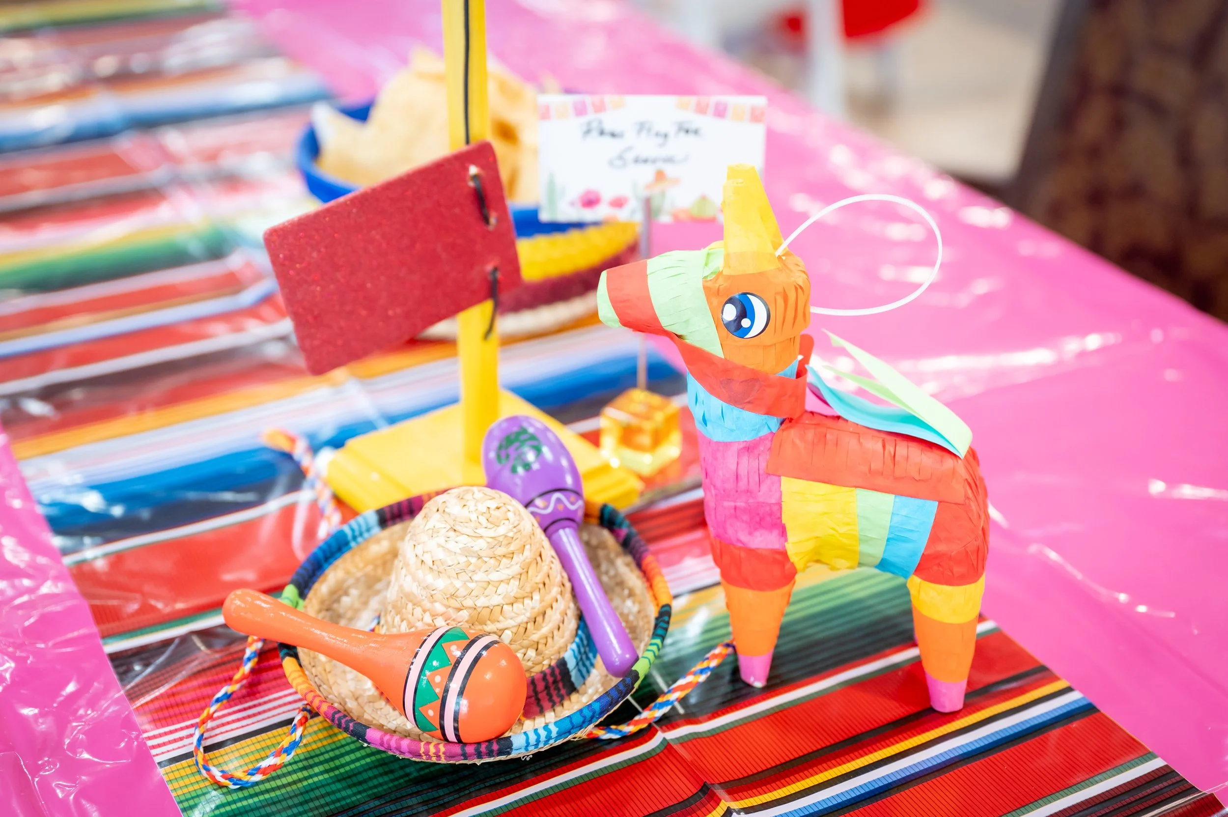 Colorful piñata in shape of a llama surrounded by pesos, a sombrero, a maraca, a purple toy microphone, and a small gift box, on a pink tablecloth at a festive celebration.