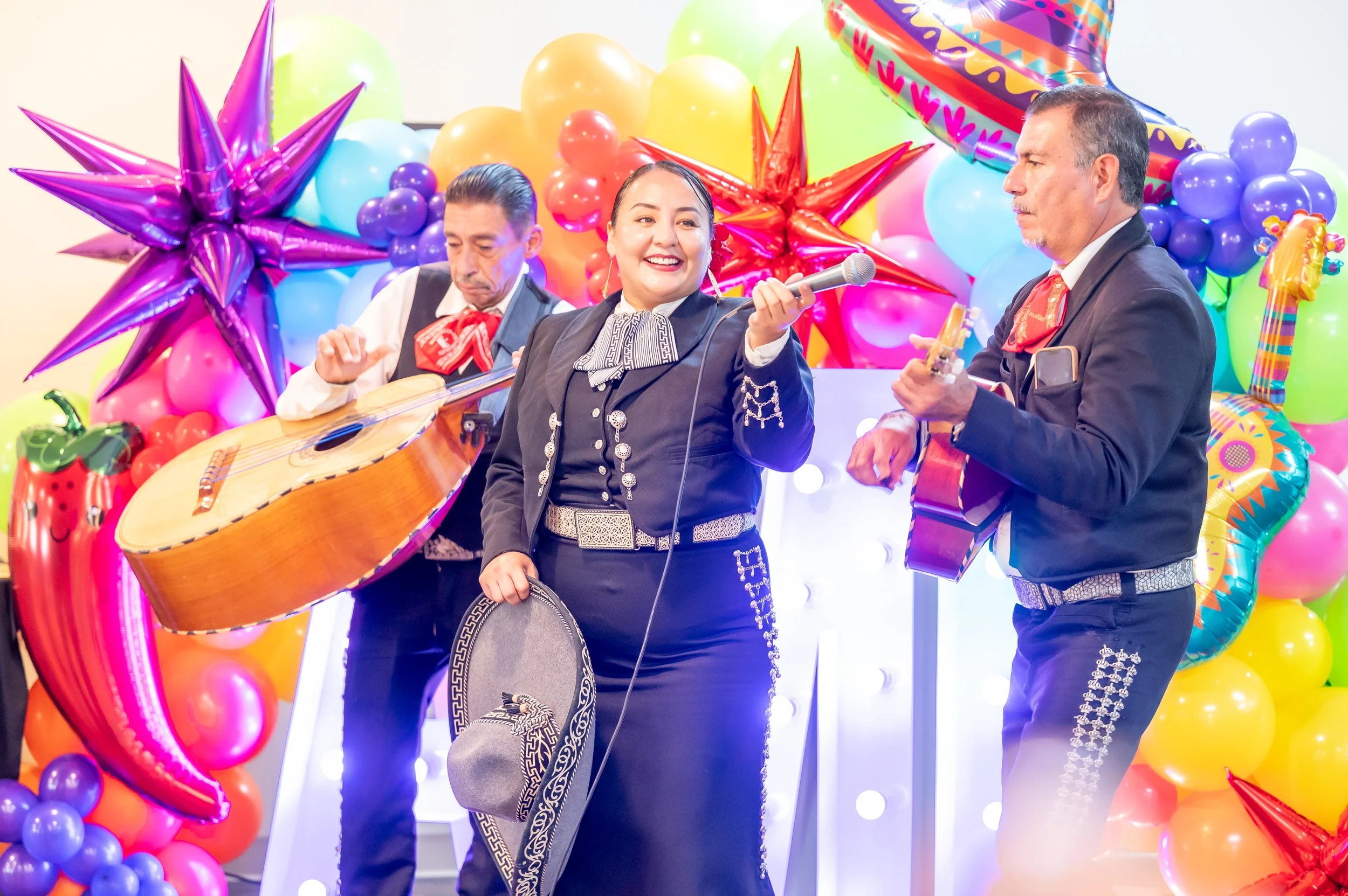 Three mariachi musicians performing in front of colorful balloons and decorations, with the woman in the center smiling while holding a microphone.