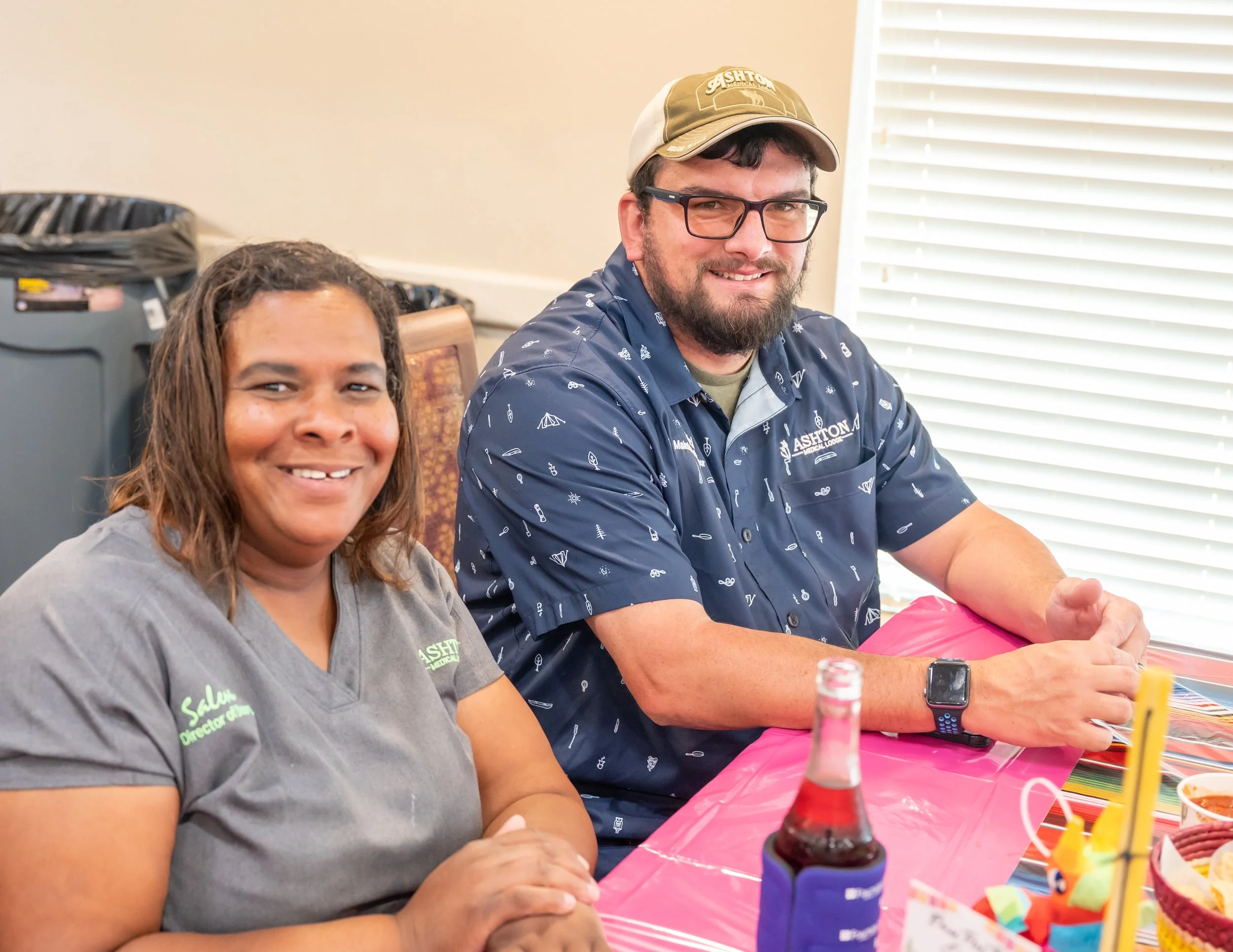 A woman and a man sitting at a decorated table, smiling. The woman wears a gray shirt with green text, and the man wears glasses, a navy shirt with white patterns, and a beige hat. The table has a bright pink tablecloth, a bottle of soda in a blue ho