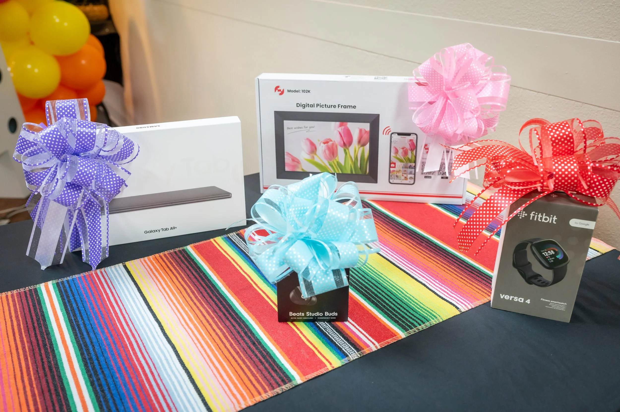 Image shows a colorful striped table runner with four wrapped gift boxes topped with large ribbons, alongside electronic gift boxes including a Samsung Galaxy Tab A9+ box, a digital picture frame box, a set of Beats Studio Buds, and a Fitbit Versa 4 