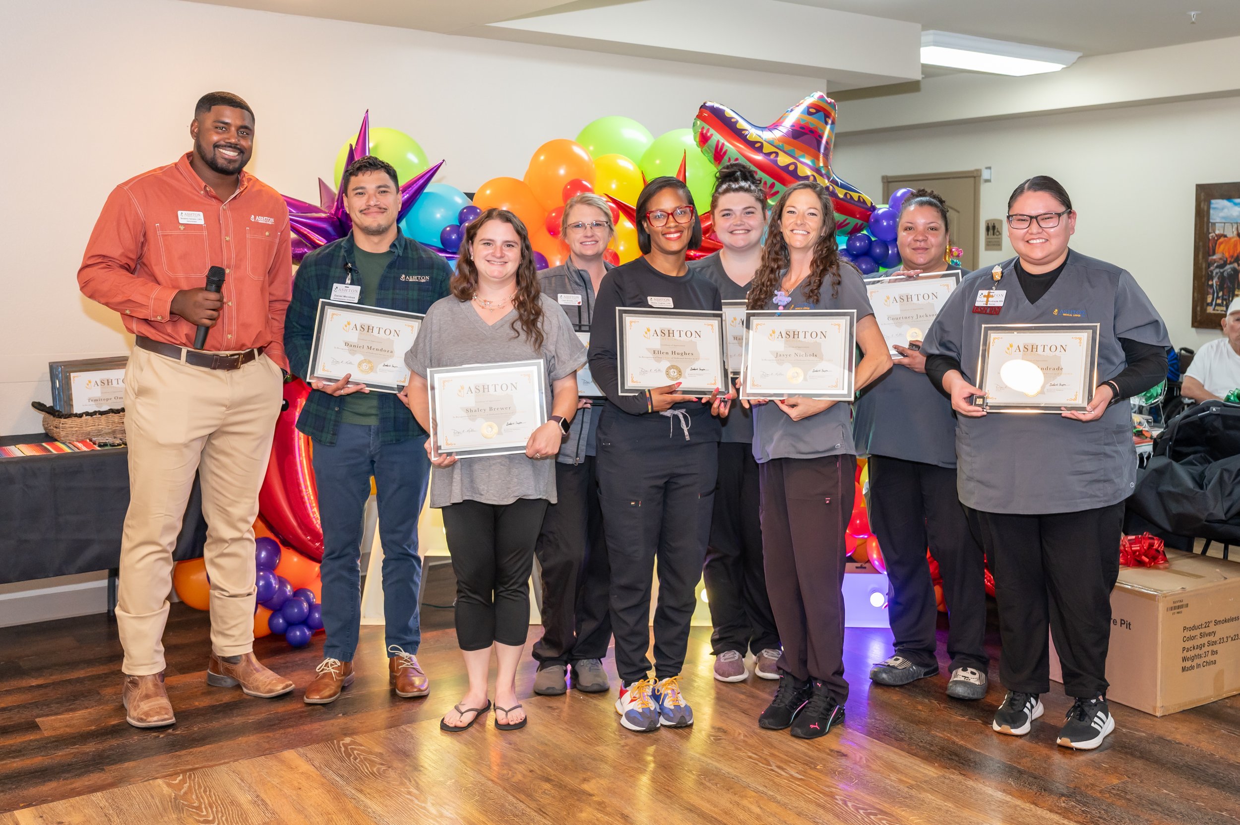 A group of nine people, including staff and award recipients, standing indoors with colorful balloons and a sombrero-shaped decoration in the background, holding certificates of achievement.