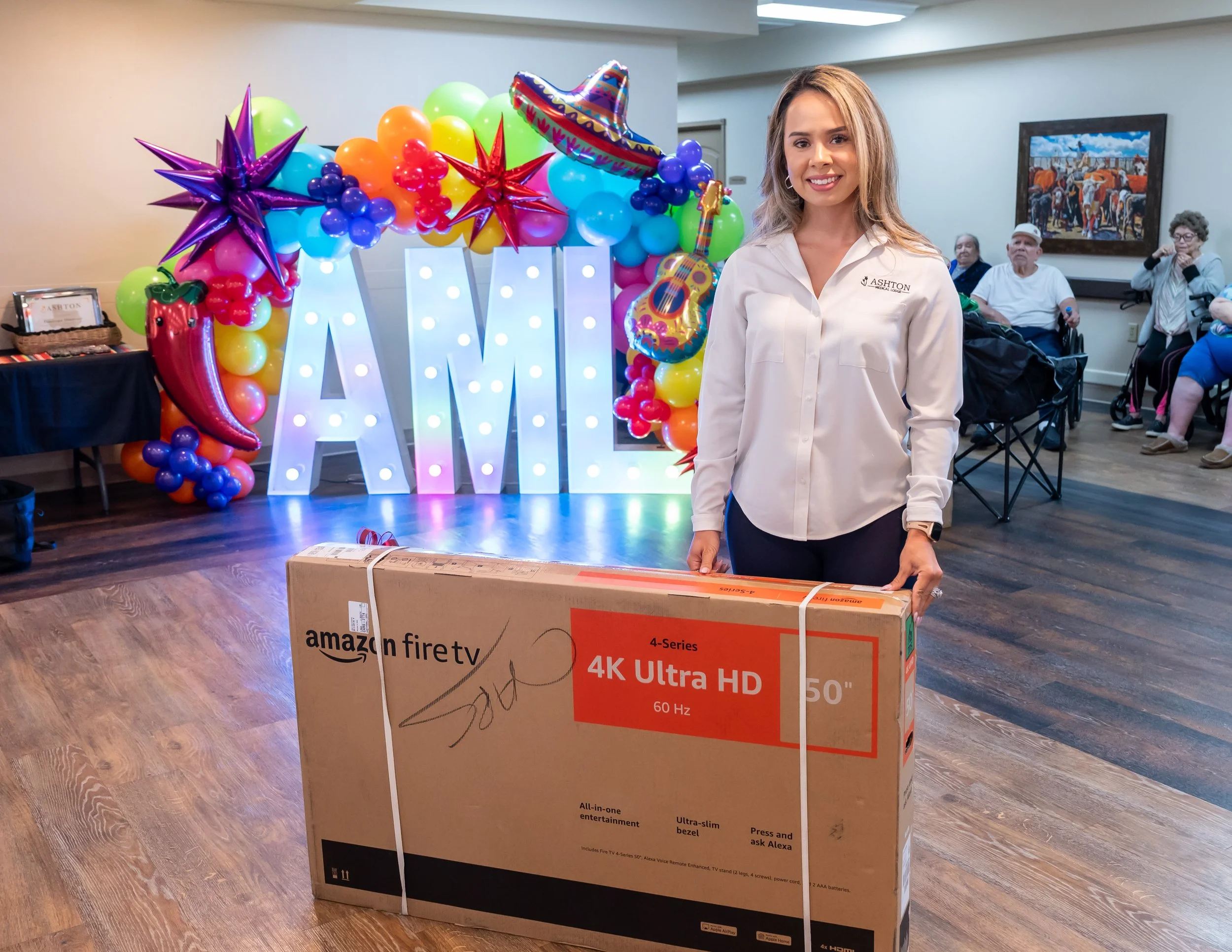 A woman standing indoors, holding a large boxed Amazon Fire TV 50-inch 4K Ultra HD television, with a colorful birthday decoration and seated guests in the background.