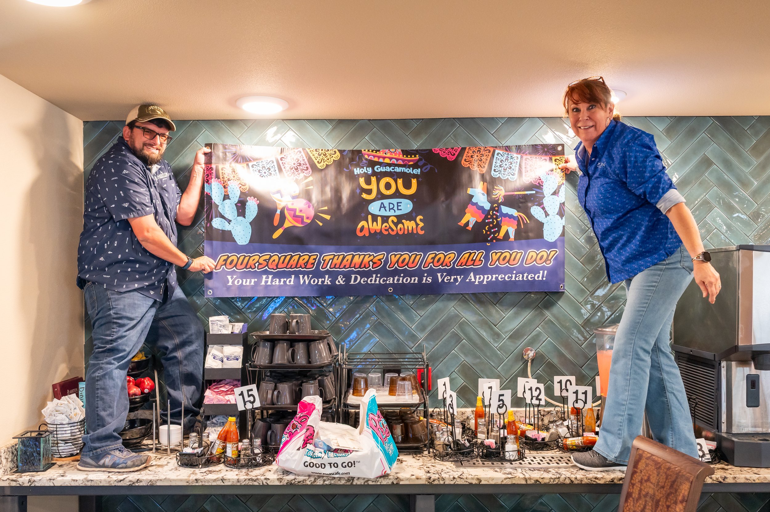 Two people standing on a table in front of a colorful banner that reads 'You Are Awesome.' They are holding the edges of the banner. The table is set up with coffee cups, bottles, and small signs with numbers, used for a Foursquare thank you event.