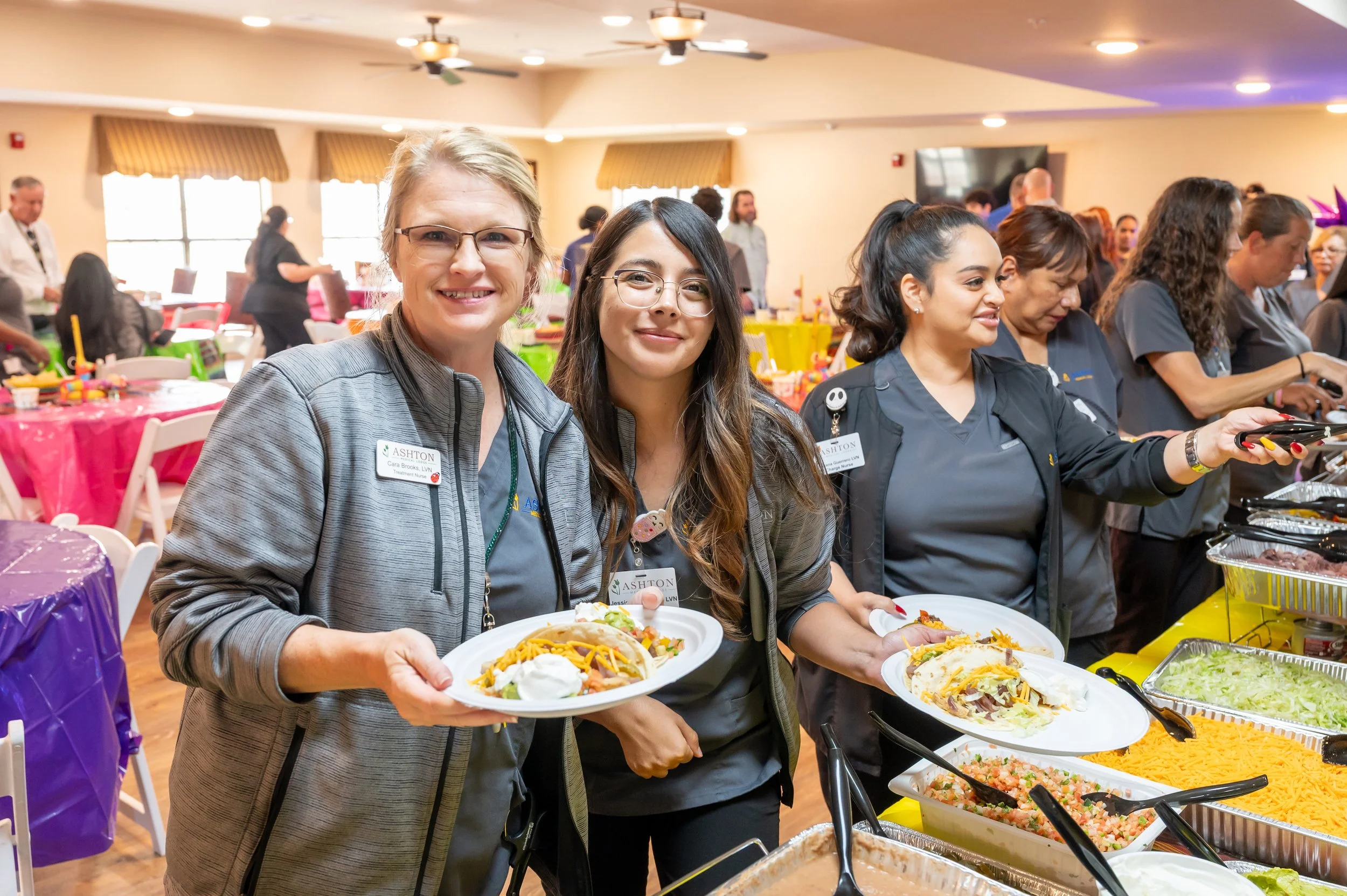 Three women in gray uniforms serving food at a buffet table during a social event, with colorful decorations and other attendees in the background.