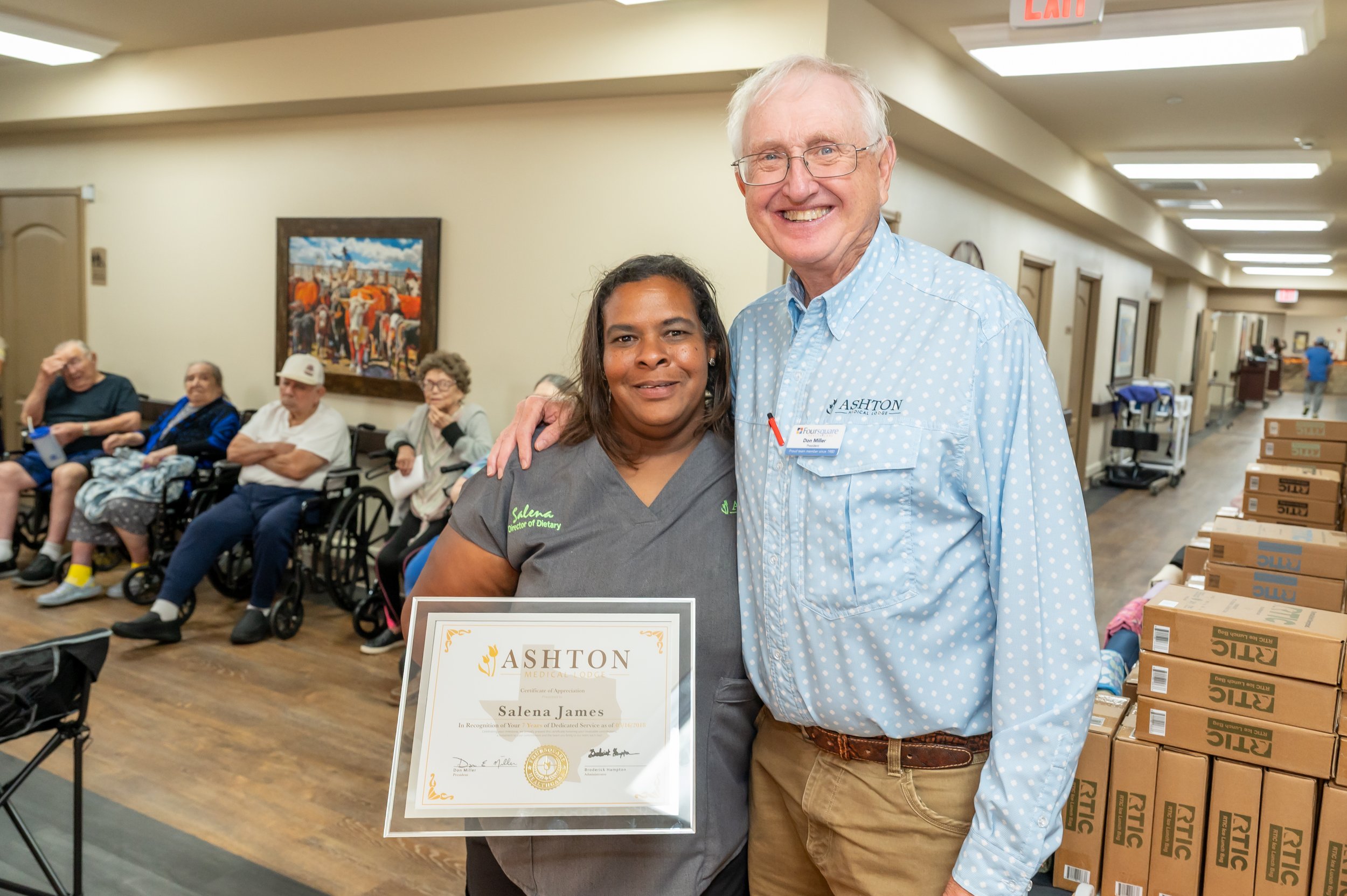 A woman holding a certificate and an older man standing beside her, both smiling for the camera. The woman is wearing medical scrubs, and the man is wearing a light blue polka dot shirt. In the background, there are several seated elderly people in a