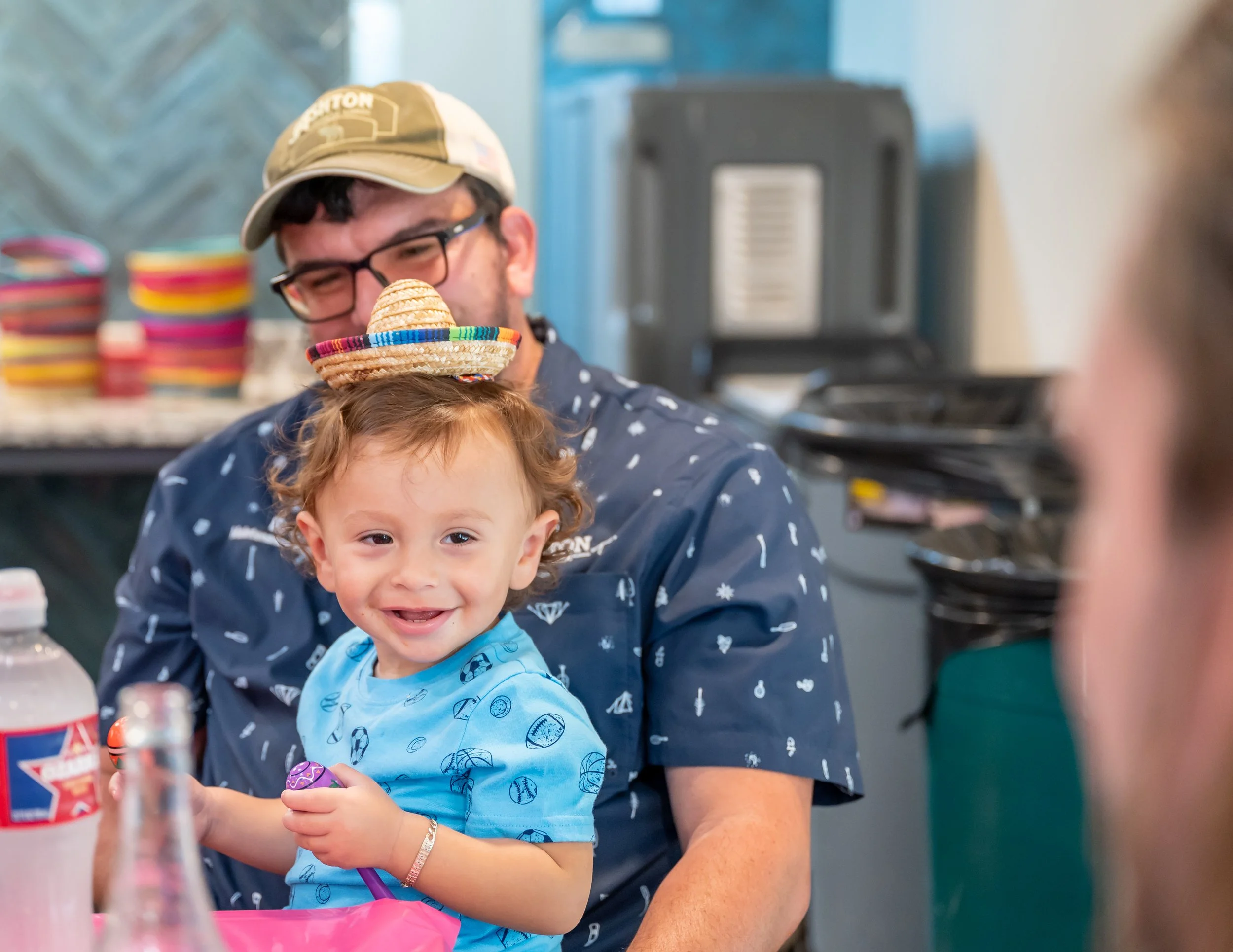A young boy with curly hair smiling at a birthday party, wearing a small sombrero hat balanced on his head, with an adult man behind him wearing glasses and a tan cap, both sitting at a table with colorful plates and drinks.