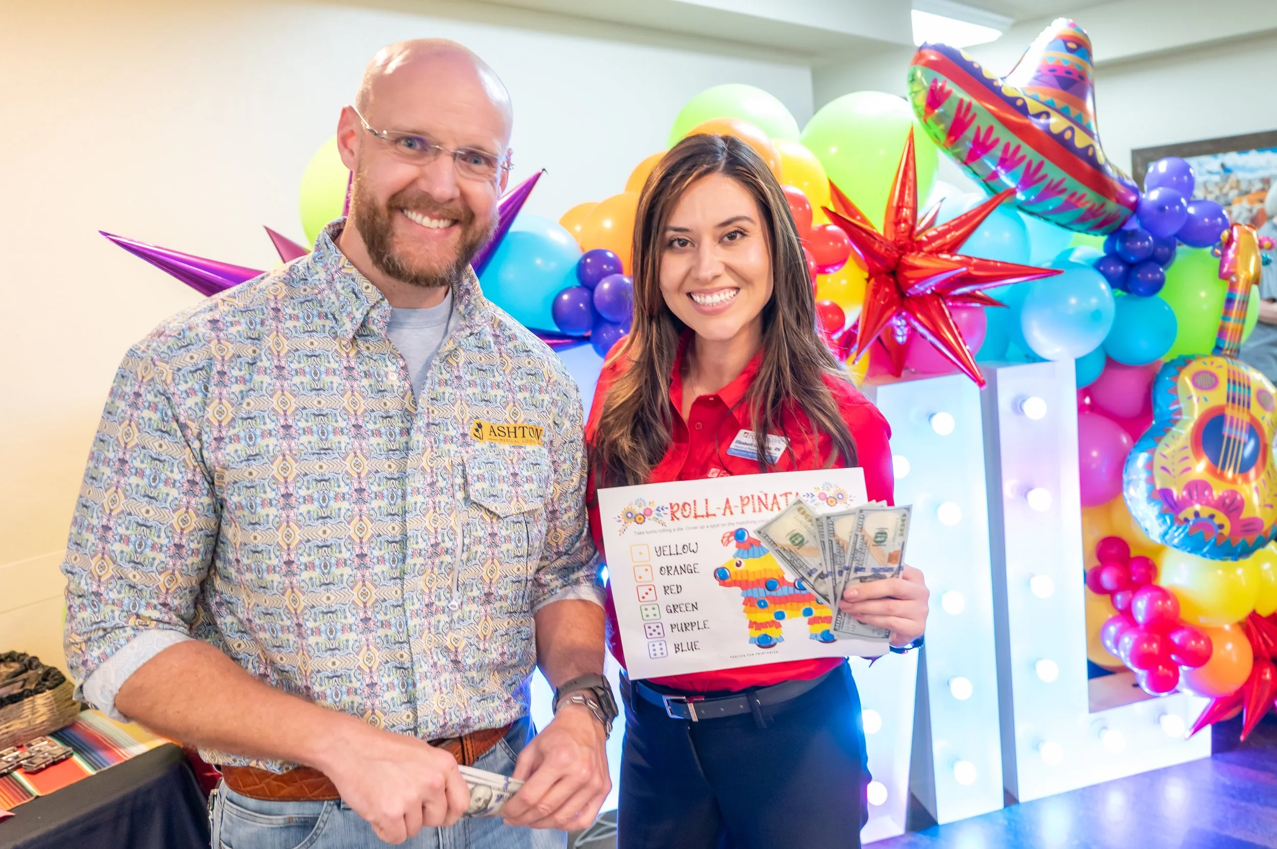 A man and a woman smiling at a celebration with colorful balloons and decorations. The woman is holding money and a game card for a game called 'Roll-A-Pinata.'