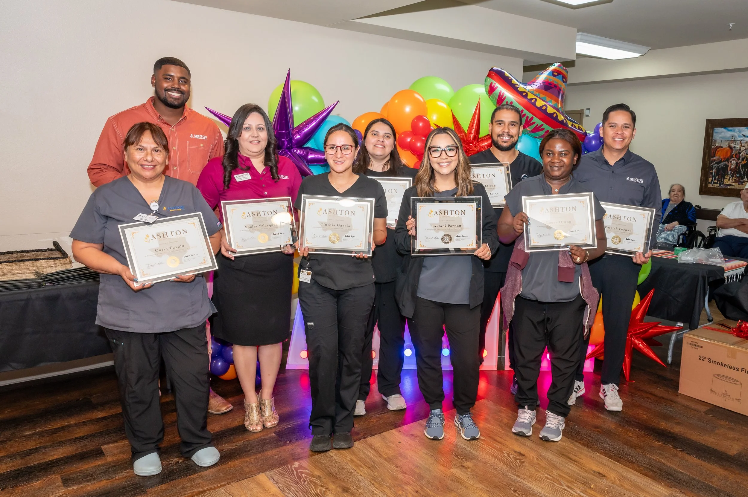 Group of nine people holding certificates, standing in front of colorful balloons and party decorations, celebrating an achievement or recognition event.