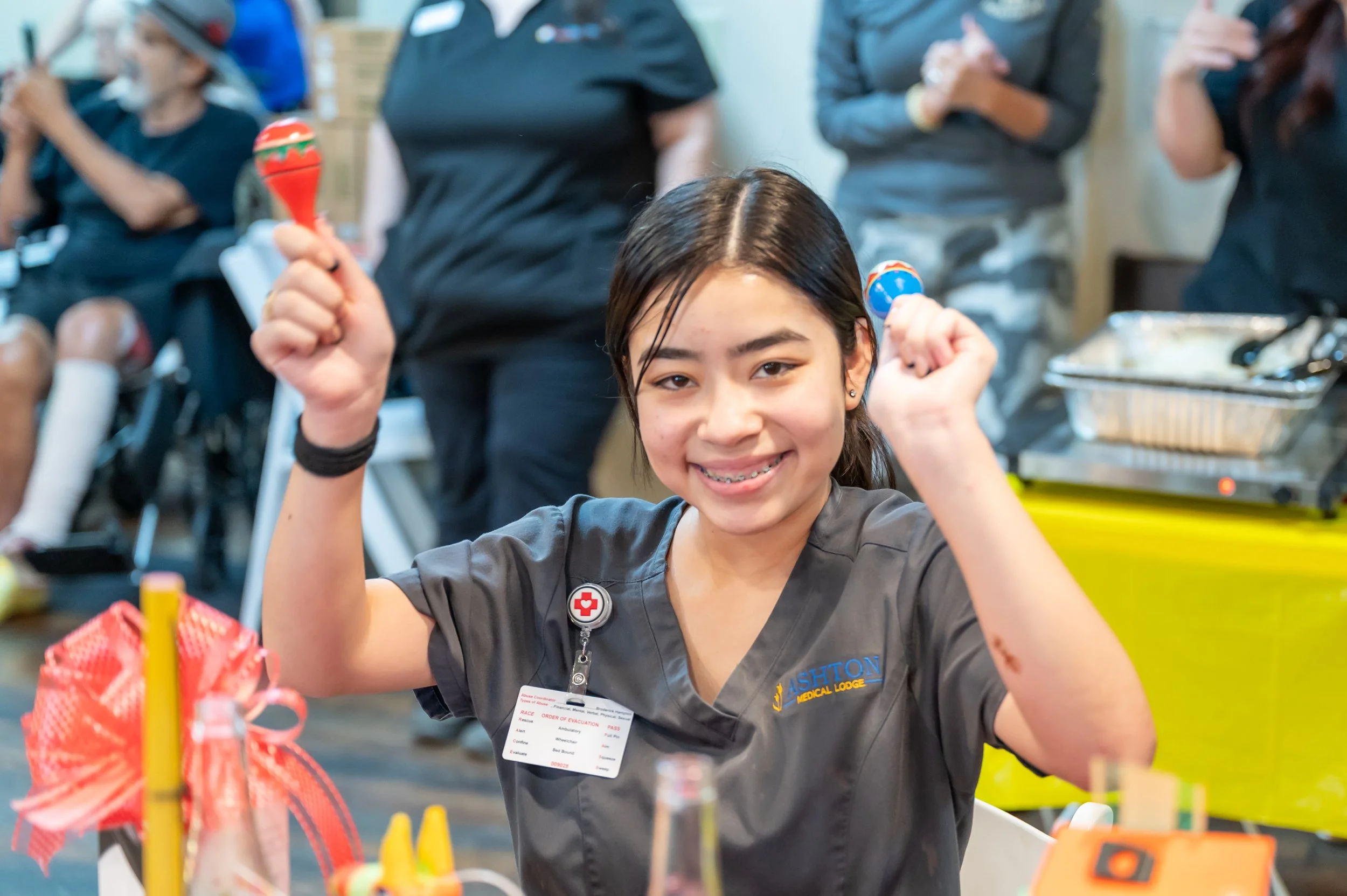 Young girl smiling with braces, wearing a gray medical uniform and a volunteer badge, holding maracas, at a community event