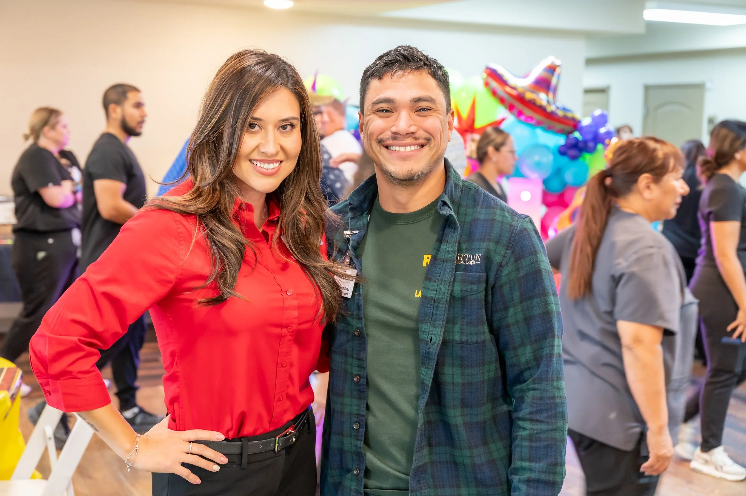 A woman and man smiling at the camera at a festive gathering with colorful balloons and people in the background.