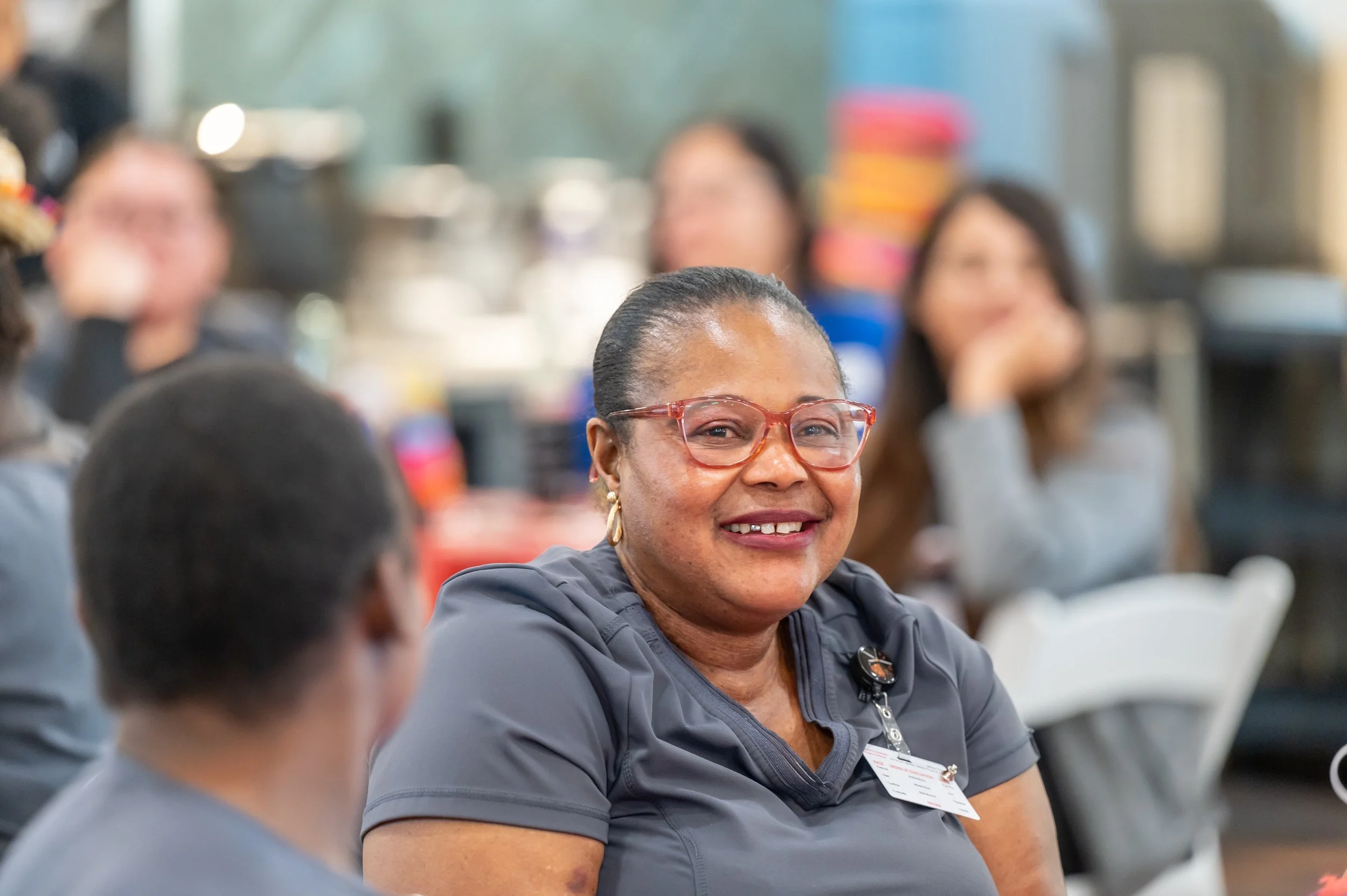 A smiling woman wearing glasses and a gray uniform, sitting at a table in a busy indoor setting with other women in the background.