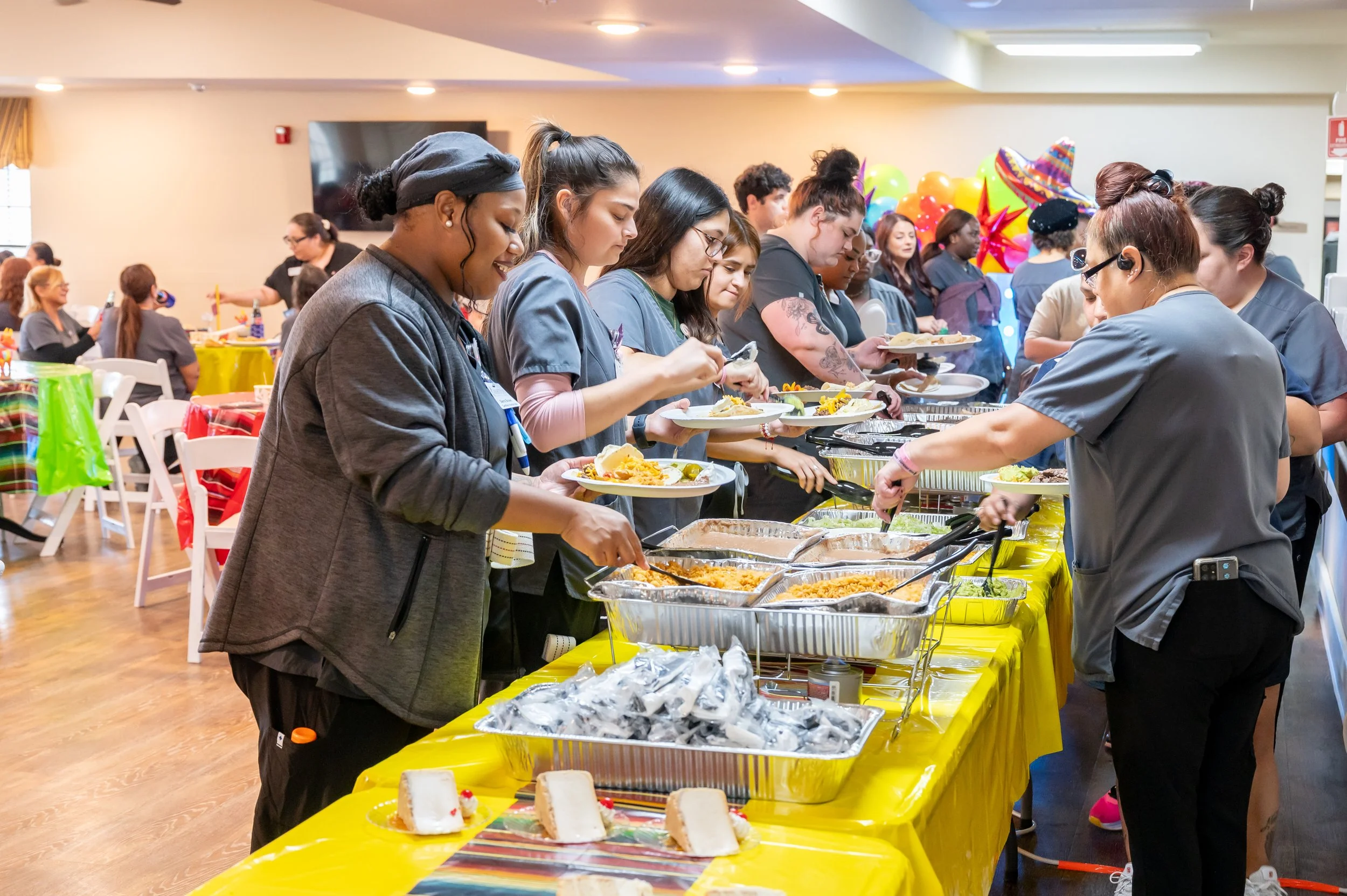 People standing in line at a buffet table serving themselves food at a festive party with balloons and decorations in the background.
