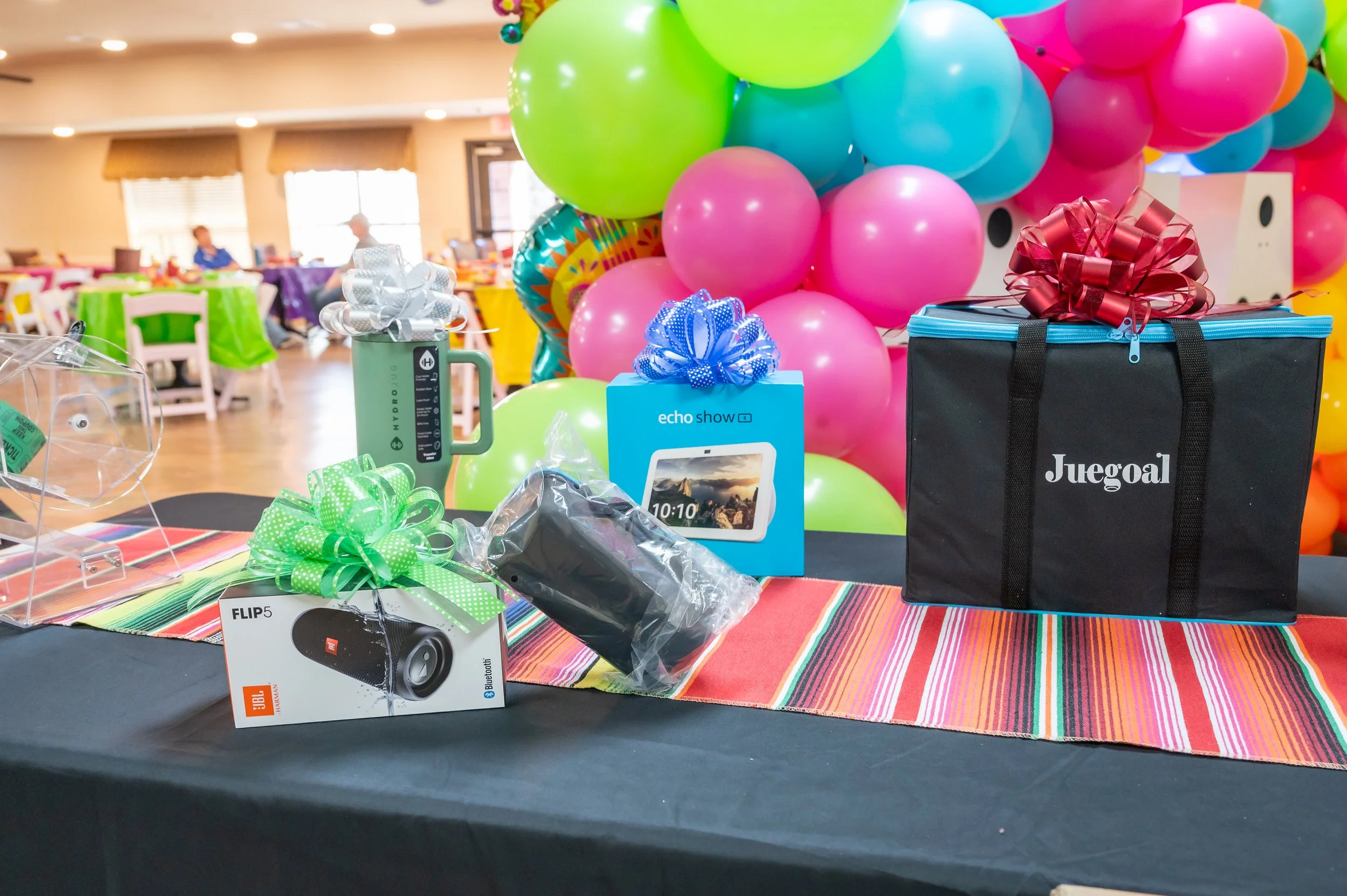 Table with gifts, including a JBL Flip 5 speaker with a green ribbon, an Echo Show box with a blue ribbon, a Juegual bag with a red ribbon, and a large black bag, with colorful balloons in the background.