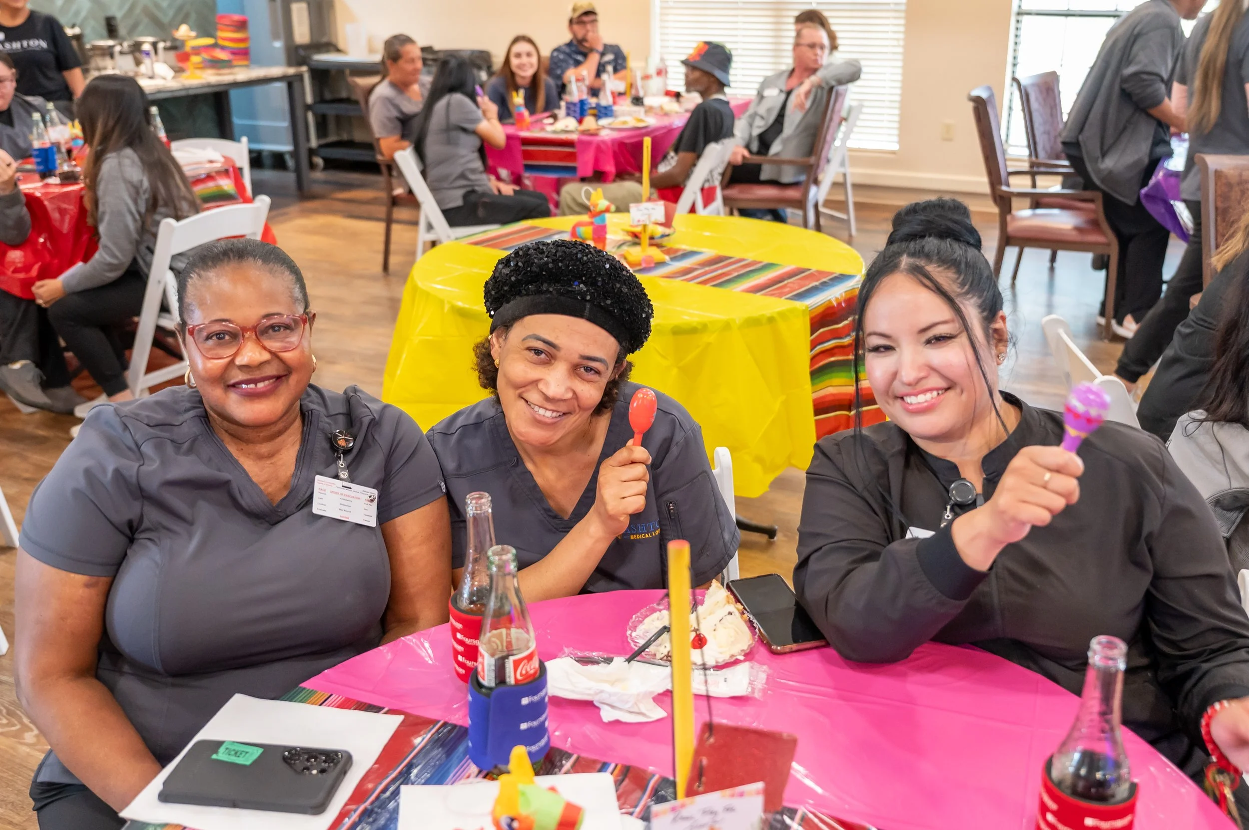 Three women smiling at a celebration table with pink and yellow decorations, holding colorful desserts, with people in the background celebrating at a party.