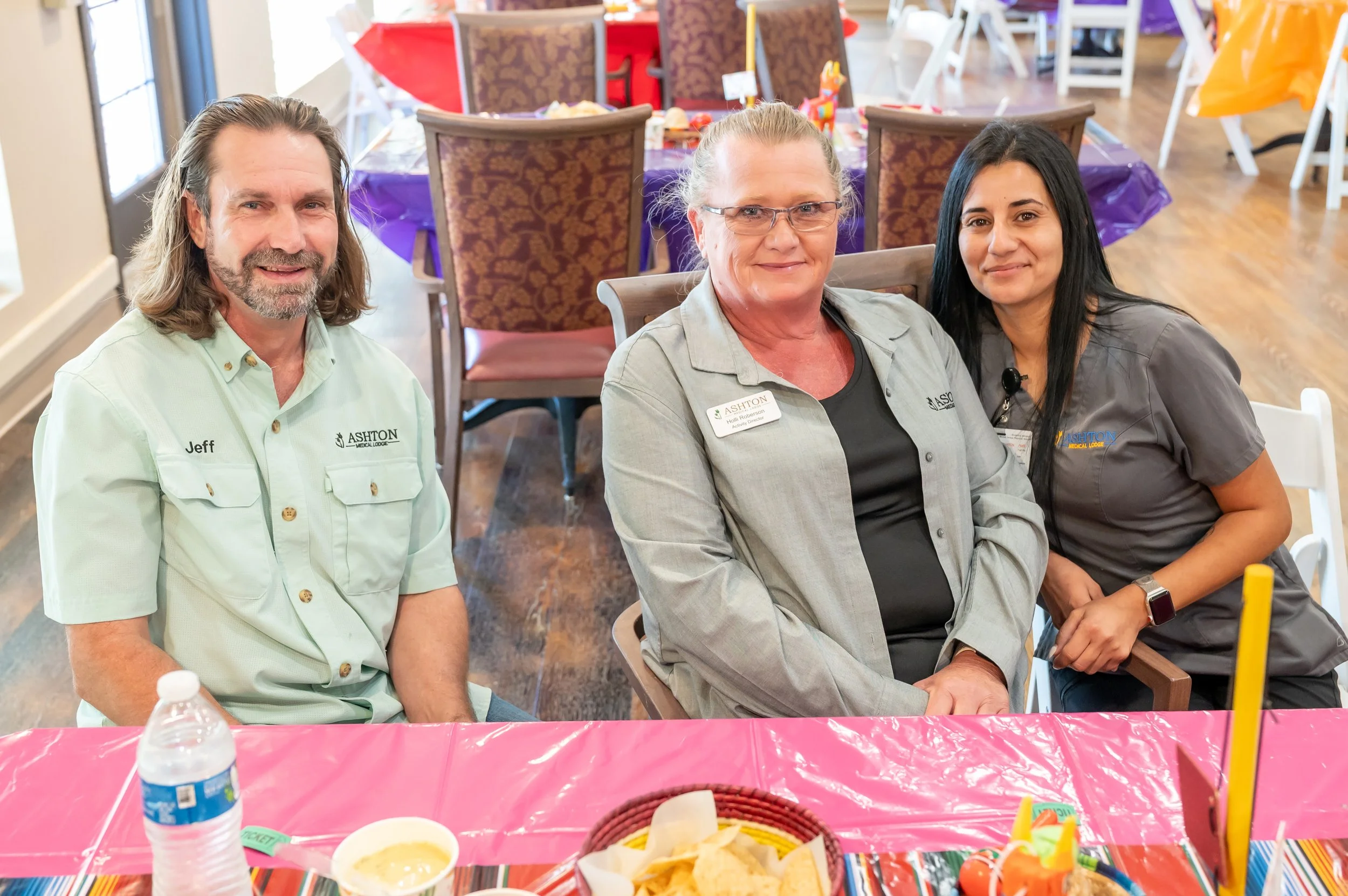Three people sit at a decorated table, smiling at the camera. The man on the left wears a light green shirt with "Jeff" on it. The woman in the middle wears a gray jacket with a name tag that reads "Ashton." The woman on the right wears gray scrubs, 