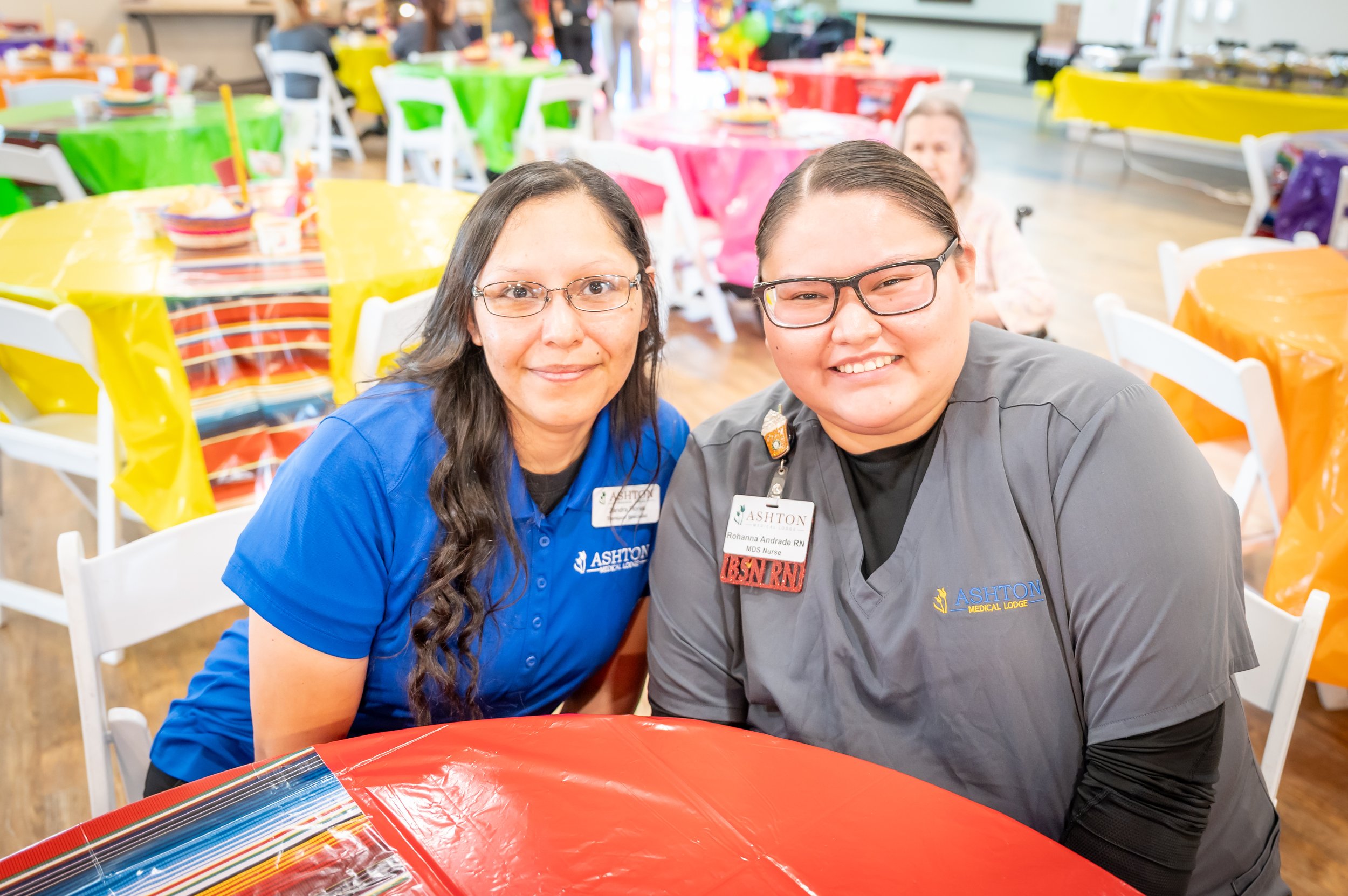 Two women wearing Ashton Medical Lodge uniforms smiling at the camera at a colorful party or event with tables covered in bright tablecloths in the background.