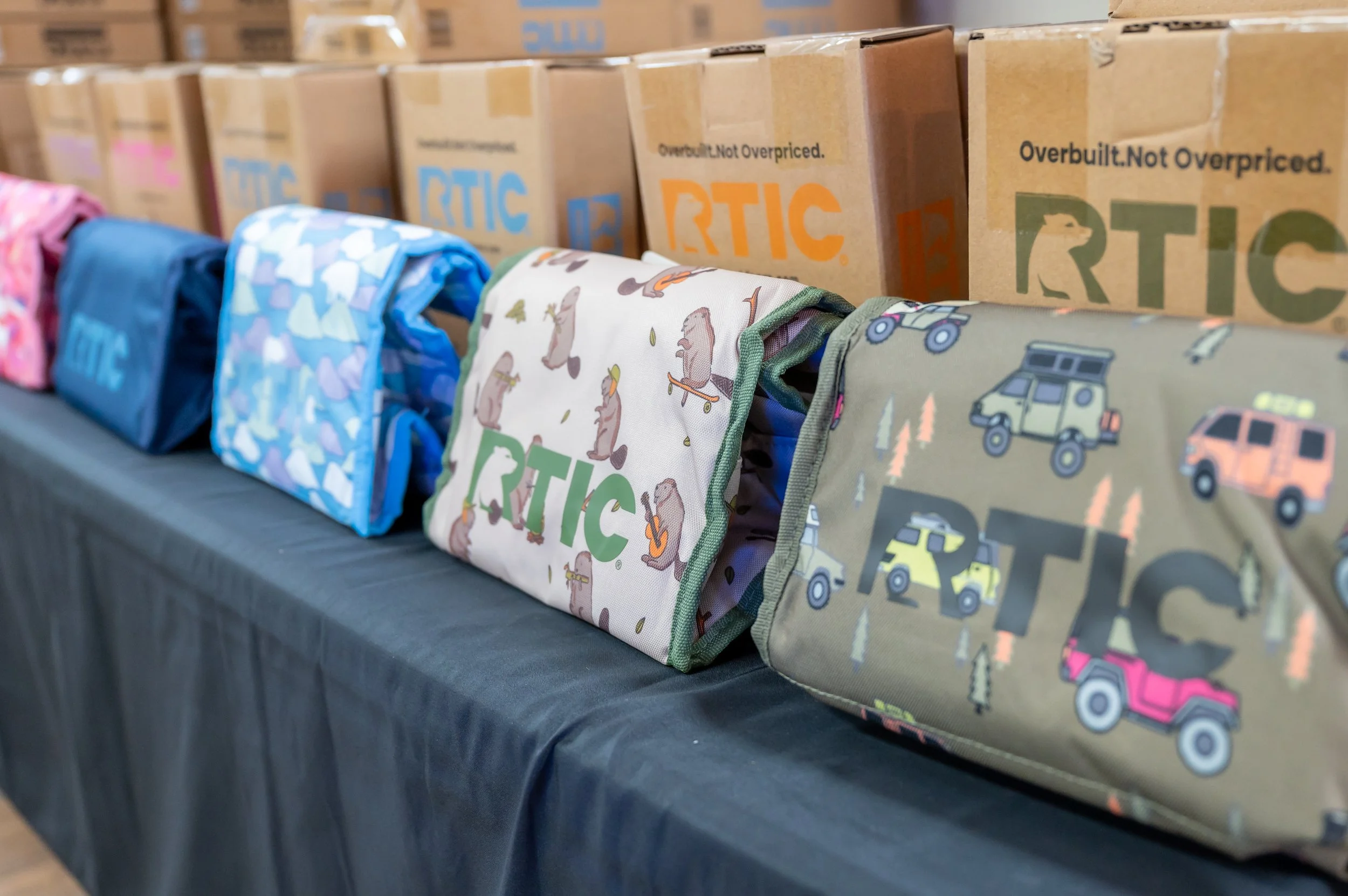 A row of cloth lunch bags with outdoors and animal prints, placed on a black table with boxes labeled 'RTIC' behind them.