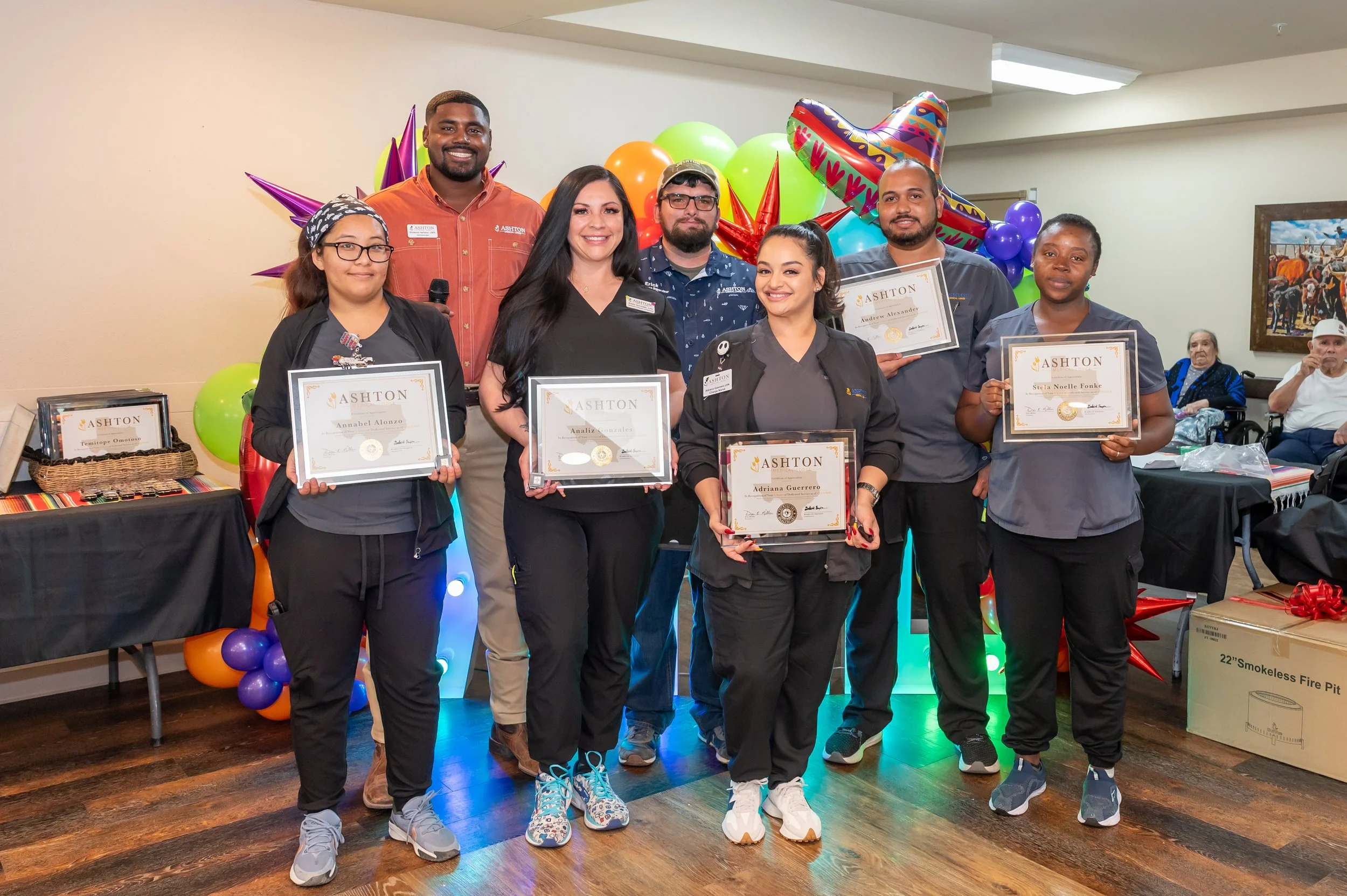 Group of seven people, five women and two men, holding certificates at a celebration event, with colorful balloons and festive decorations in the background.
