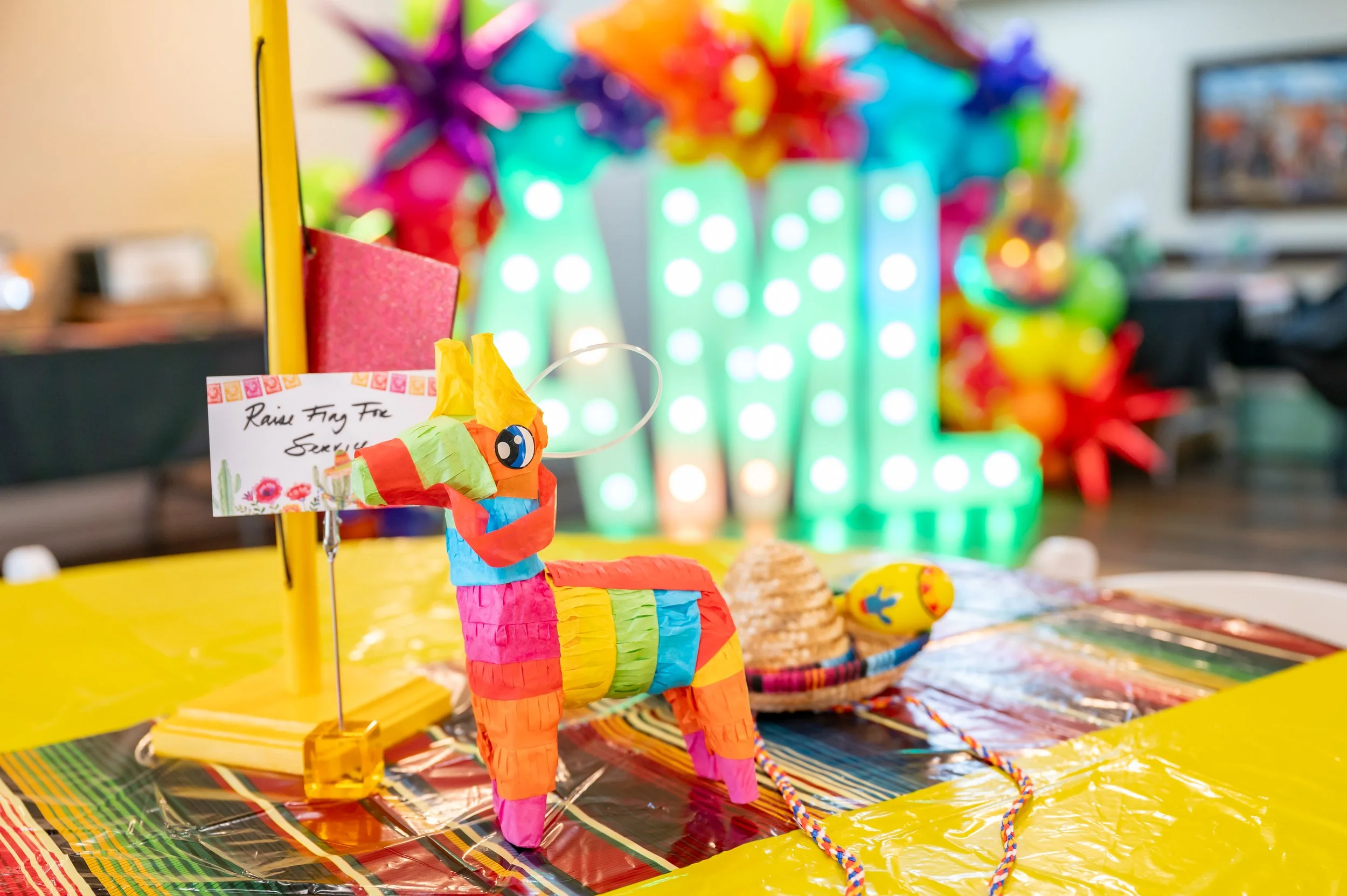 Colorful piñata with a small handwritten sign that reads 'Raise Fiy Fie Sorry,' decorated on a table with bright yellow and multicolored tablecloths, with a background of vibrant decorations and illuminated letter lights spelling out 'HAW'.