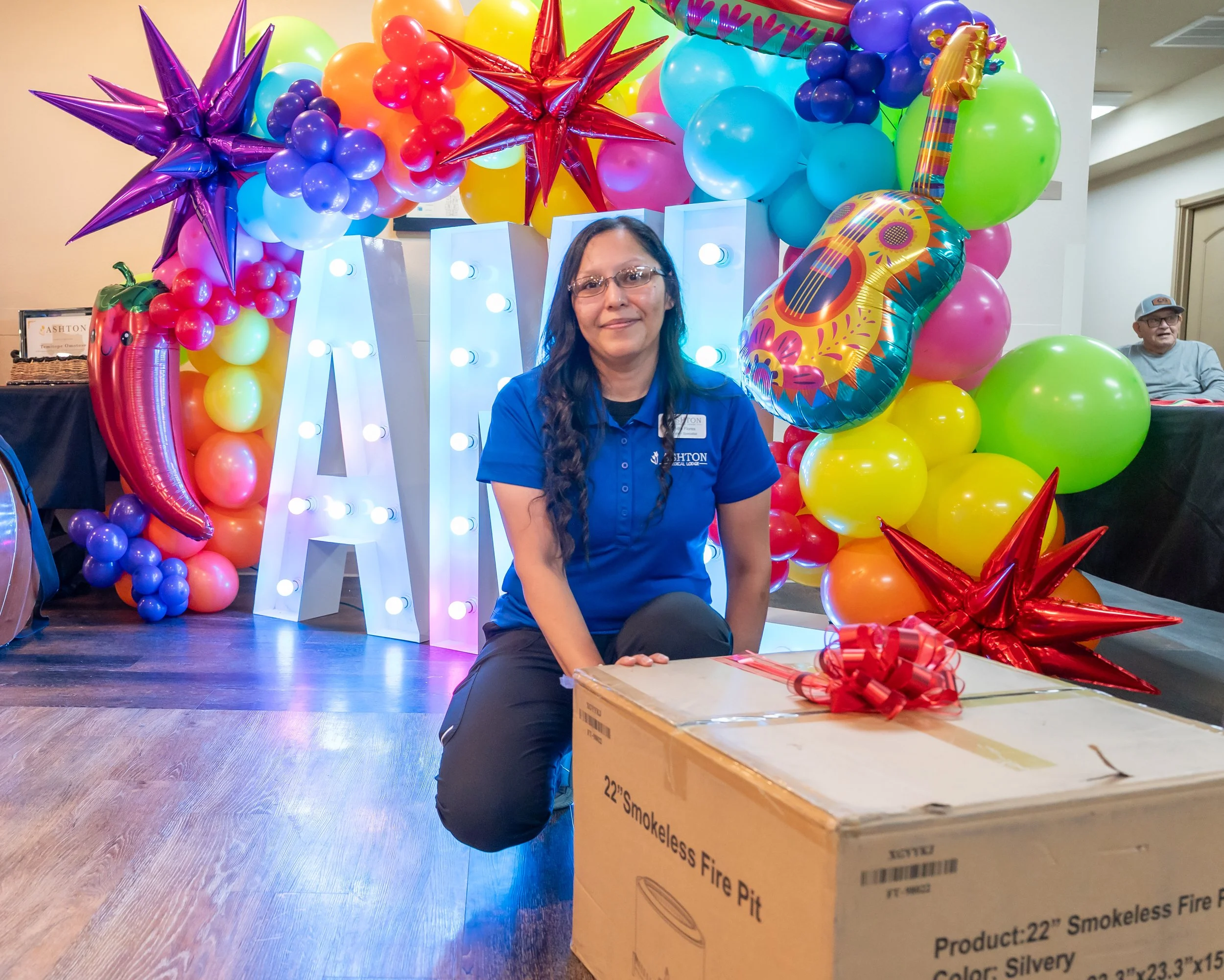 A woman kneeling in front of a colorful balloon display with large white letters spelling 'NAY'. The display includes balloons shaped like stars, a chili pepper, a guitar, and other colorful balloons. The woman is wearing glasses and a blue shirt, an