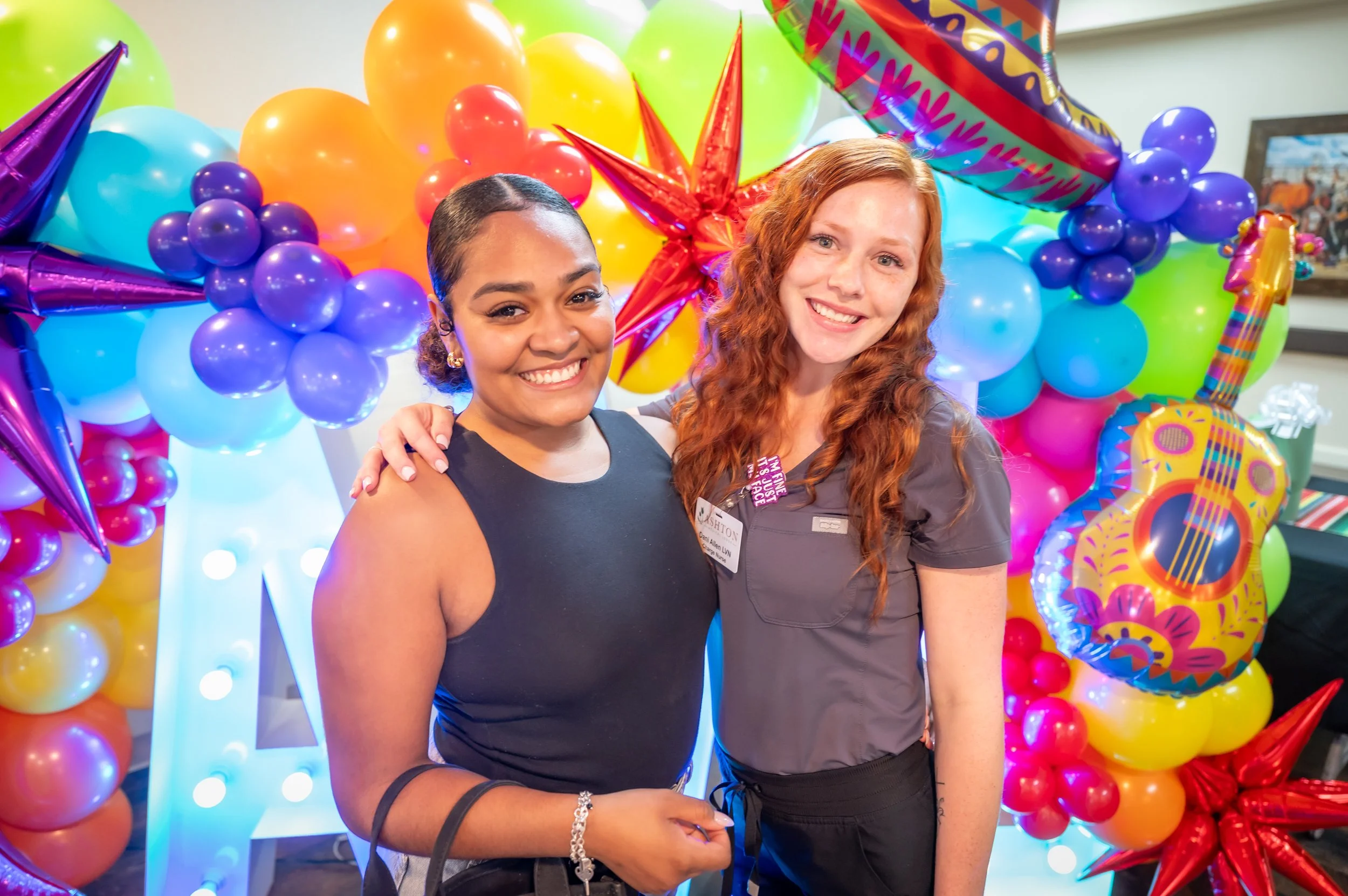 Two women smiling at a festive event with colorful balloons and decorations in the background.