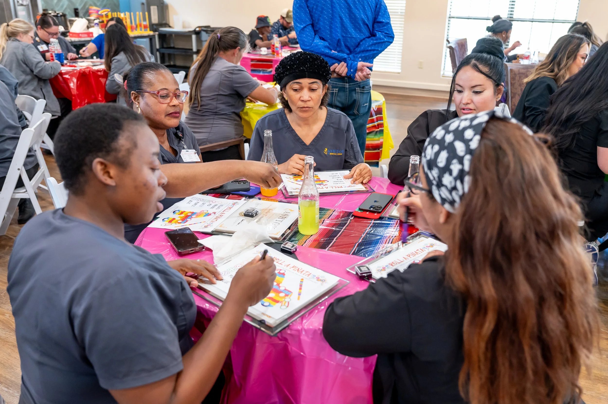 A group of women sitting around a table engaged in a discussion, with colorful decorations and various items on the table, in a bright room with large windows.