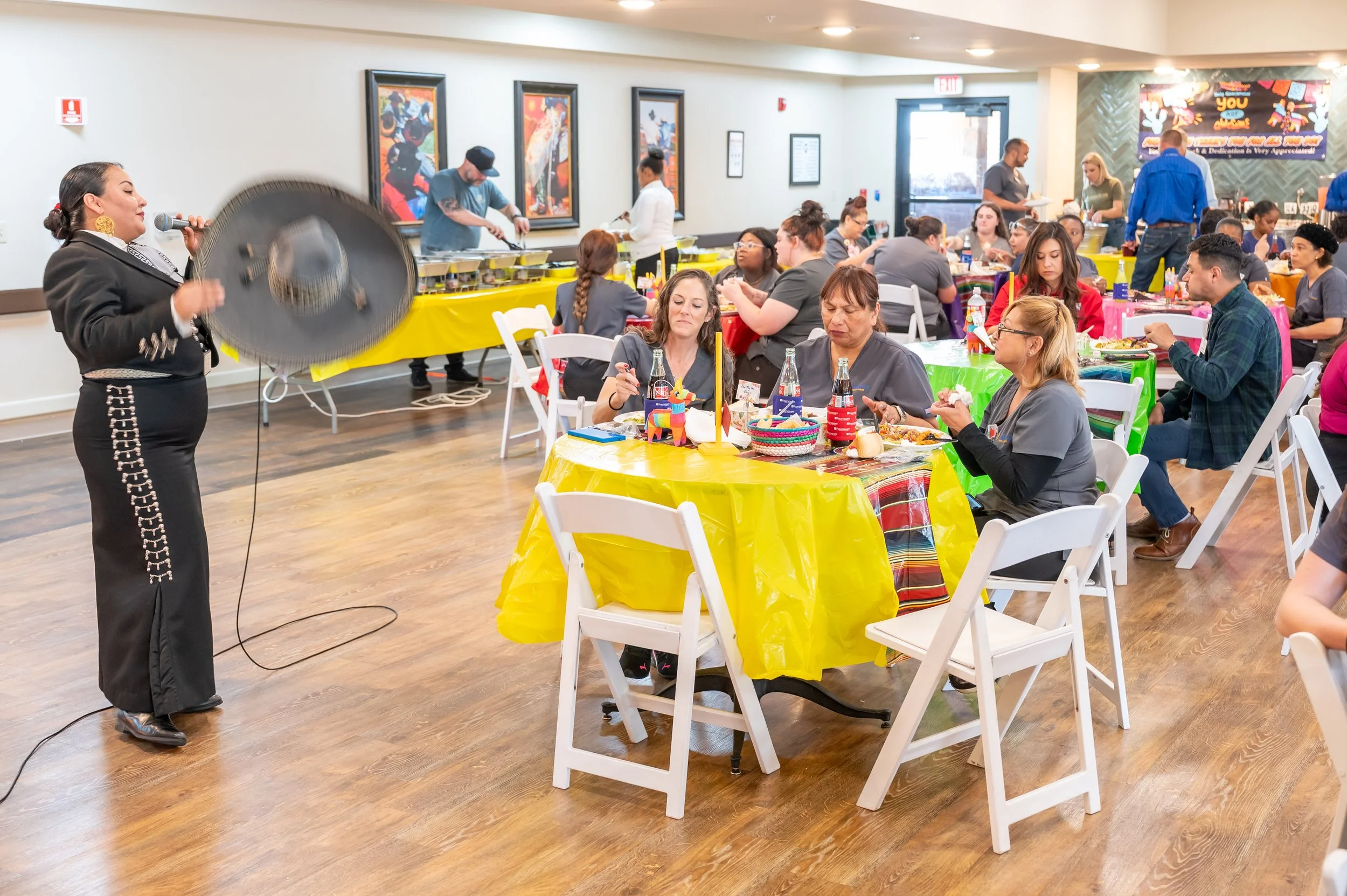 A woman singing into a microphone at a lively celebration with many guests seated at colorful tables, some eating and drinking, in an indoor event space decorated for a festive occasion