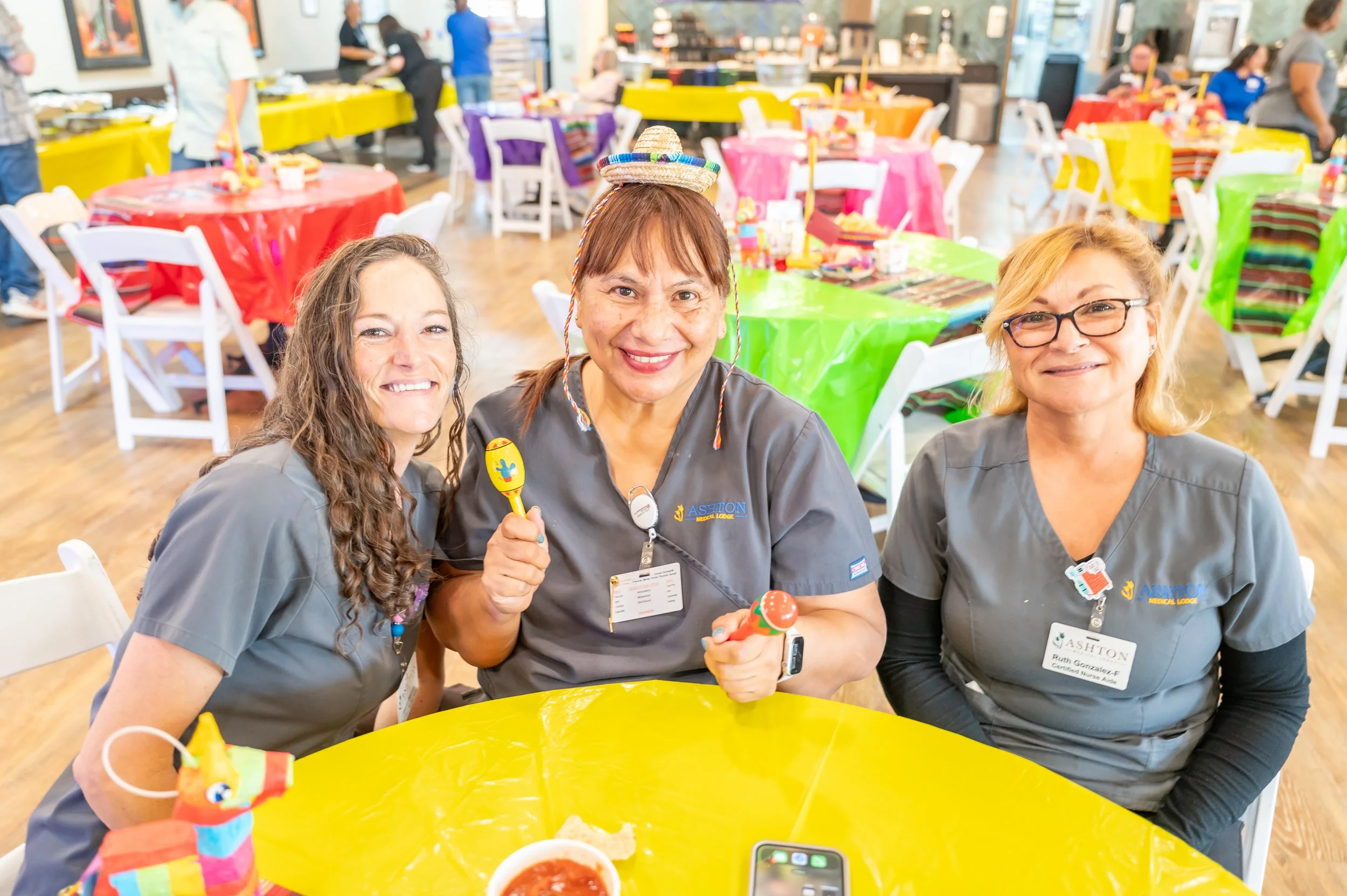 Three smiling women in gray uniforms at a festive gathering, sitting at a yellow table with colorful tablecloths and decorations, some holding toys, with more tables and people in the background.