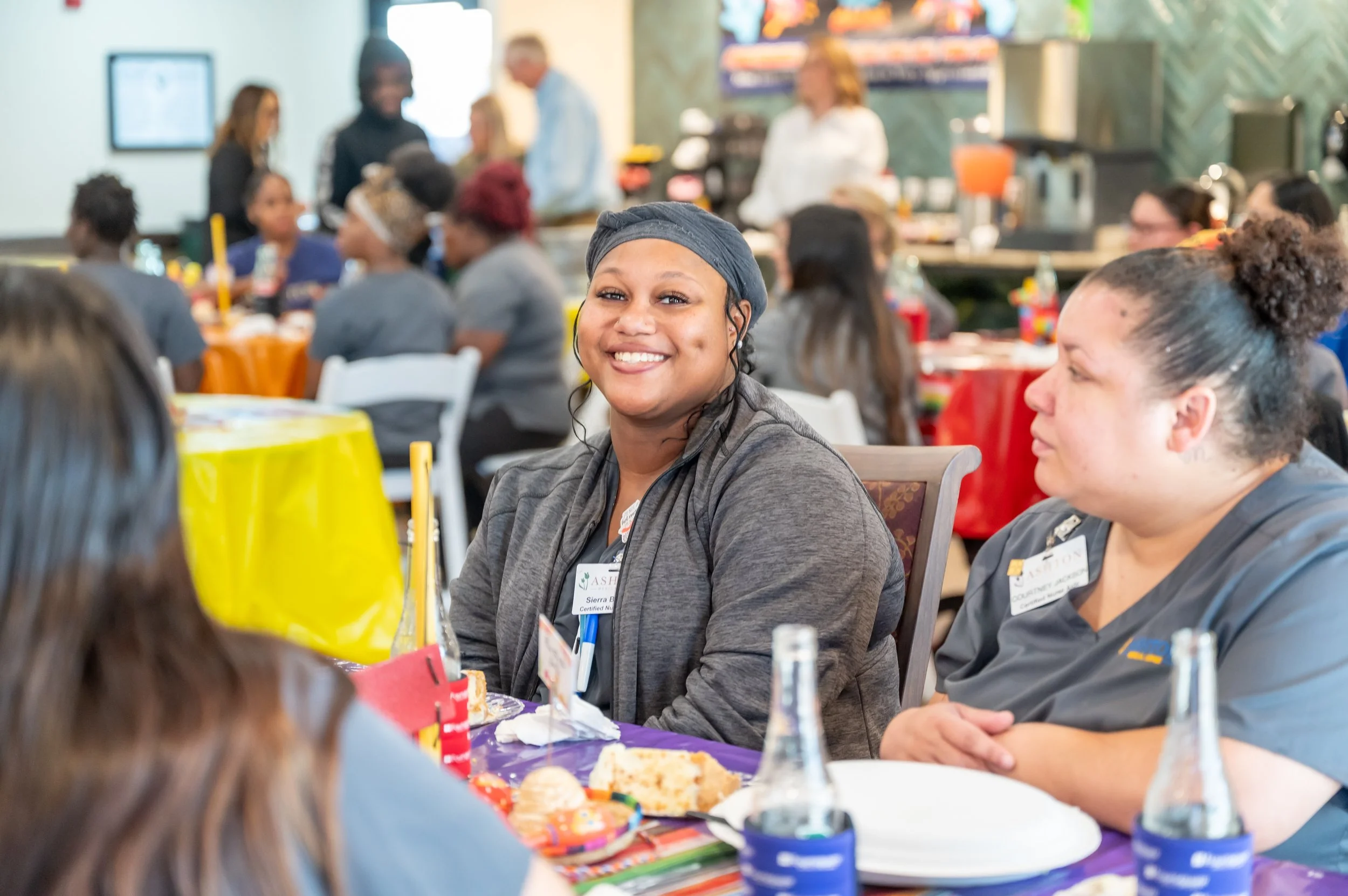 Smiling woman sitting at a table with other people at a celebration or gathering, with colorful decorations and cake on the table.